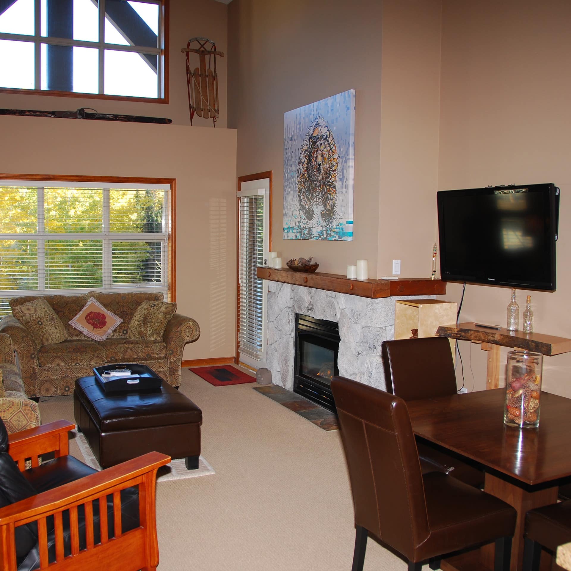 Living area with tall ceiling, tan walls, large windows, comfy furniture, wooden dining suite, TV and stone fireplace