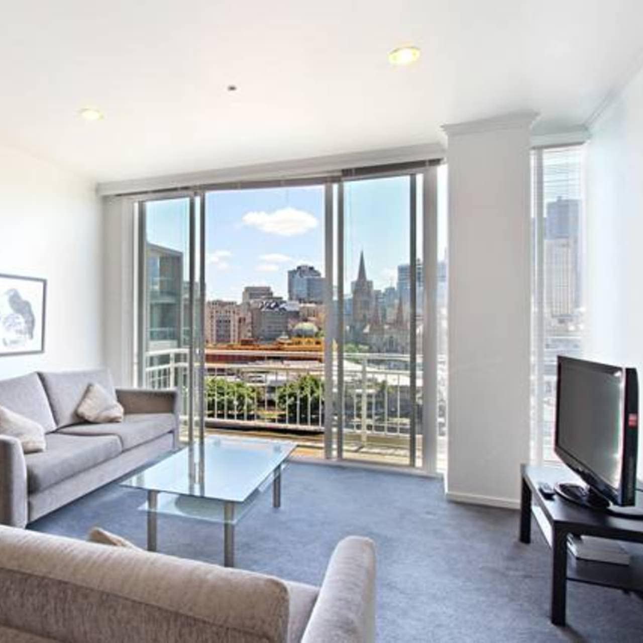 Living room with tan sofas, glass coffee table, flat-screen TV and glass sliding doors to attached balcony with city views.