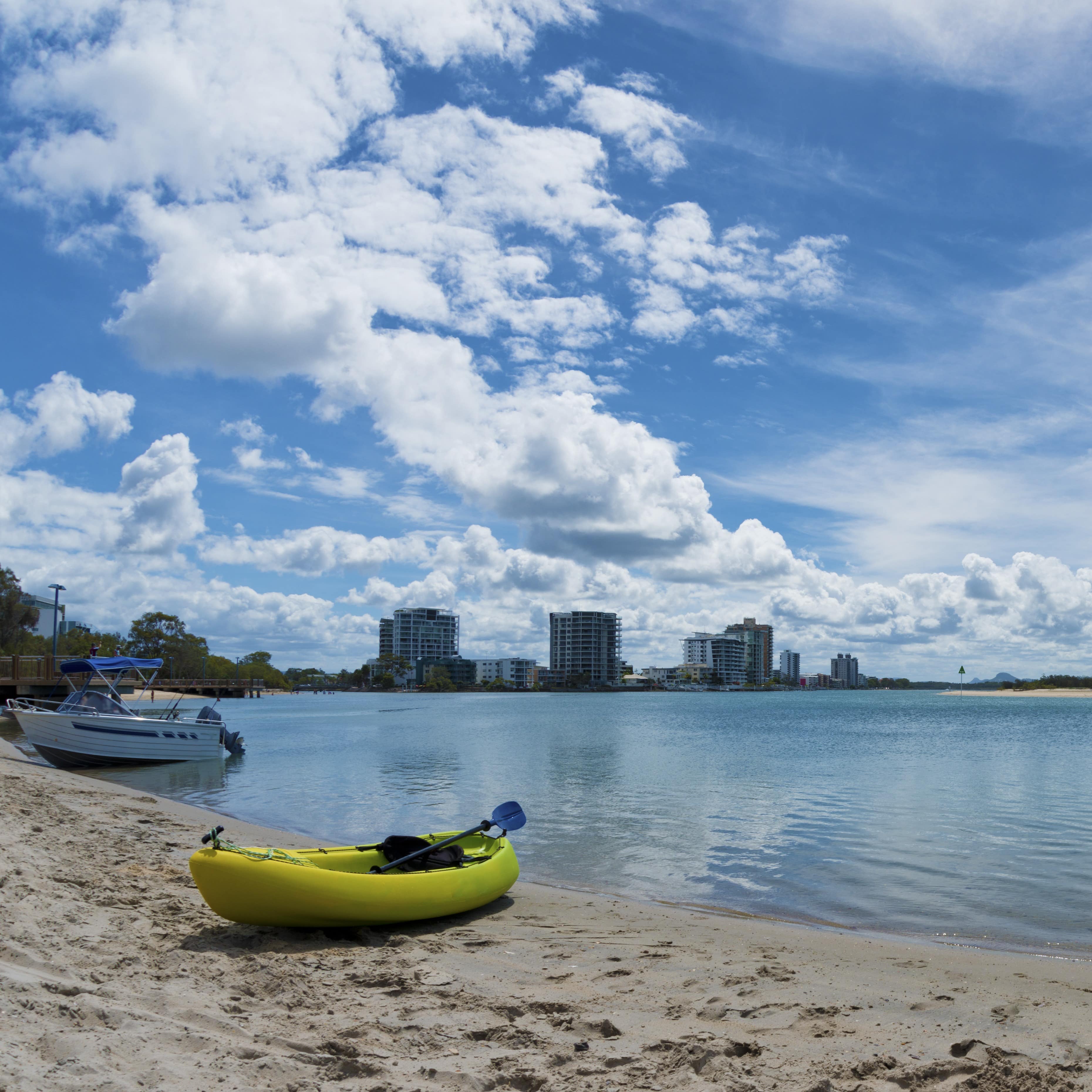 Cotton Tree Maroochy River scene with kayak, boat on shore and city in background