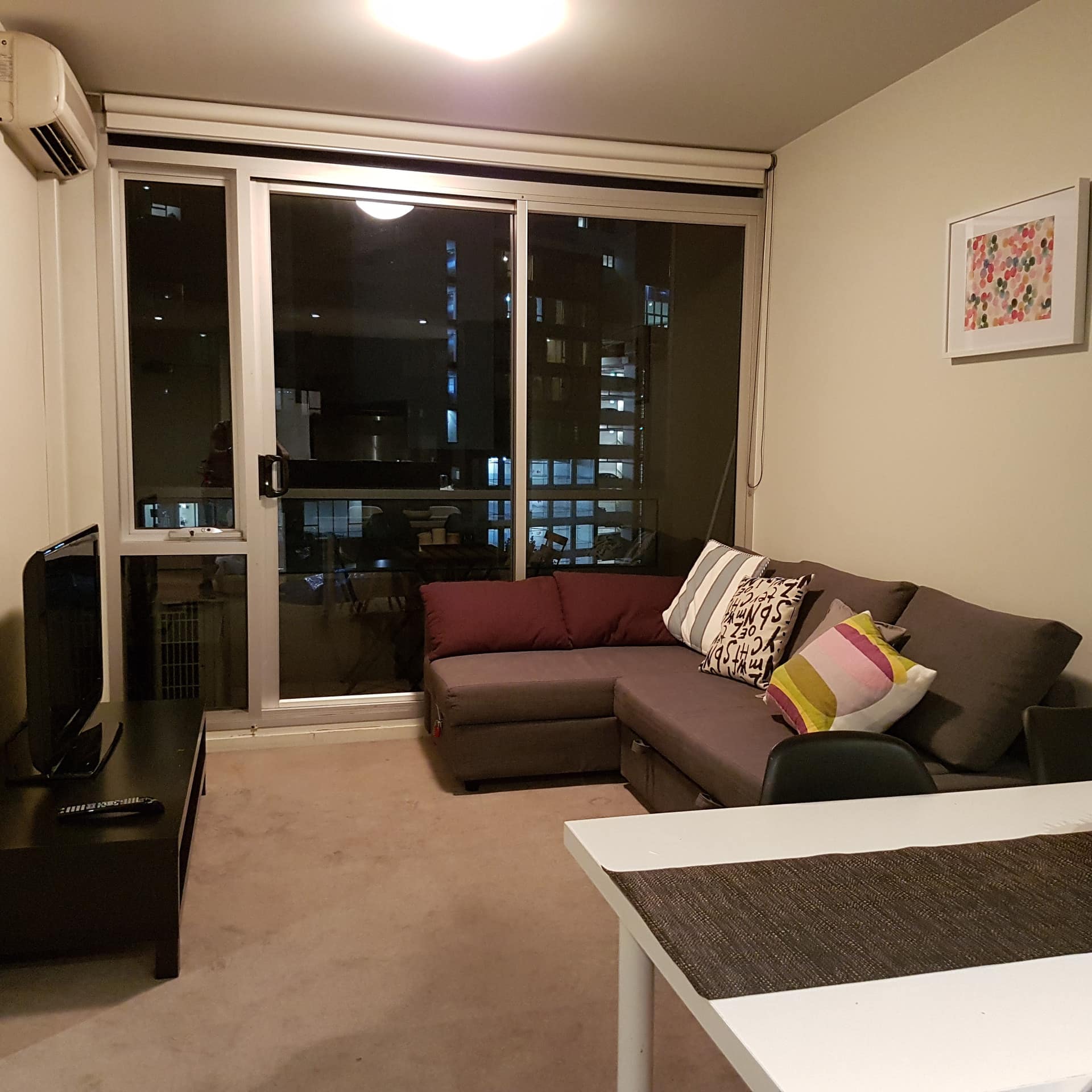 A living room with beige carpet, a brown sectional, flatscreen TV, sliding glass doors to attached balcony with city views.