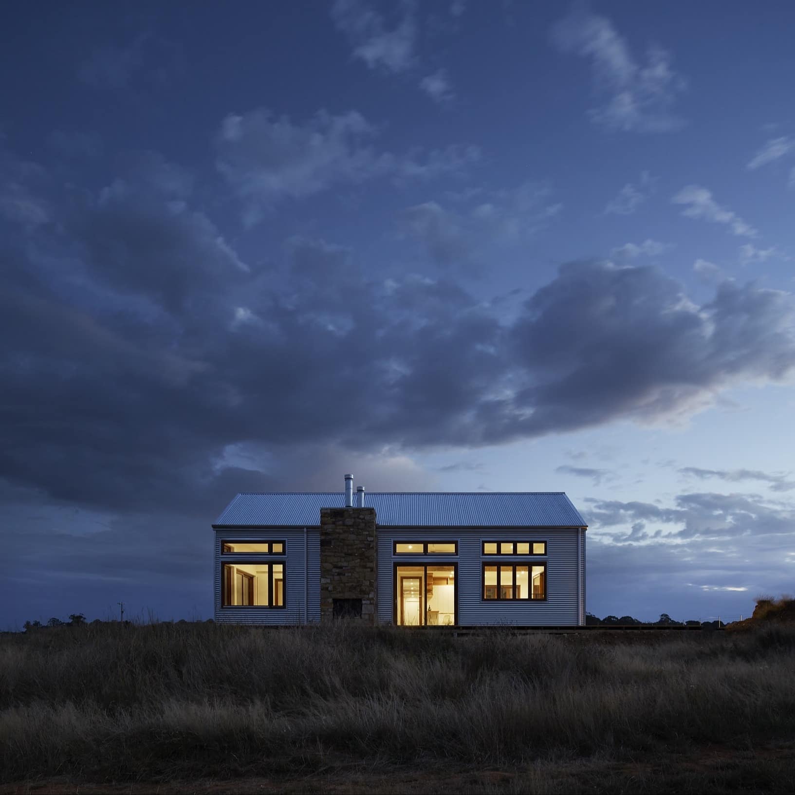 White cottage with lots of windows and a stone fireplace on farmland at dusk