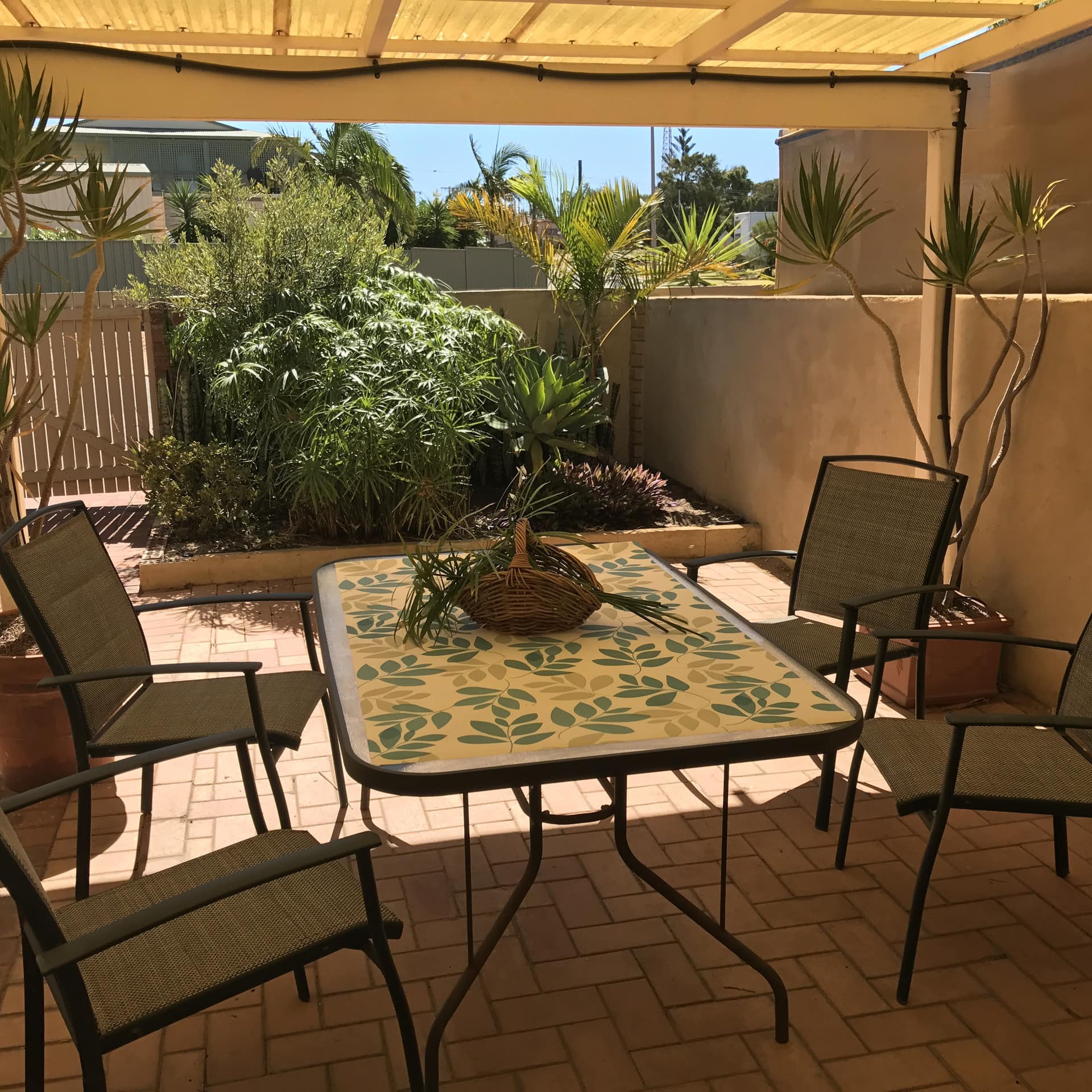 A covered patio with a black metal dining set, potted plants, and a plant bed with tropical plants.
