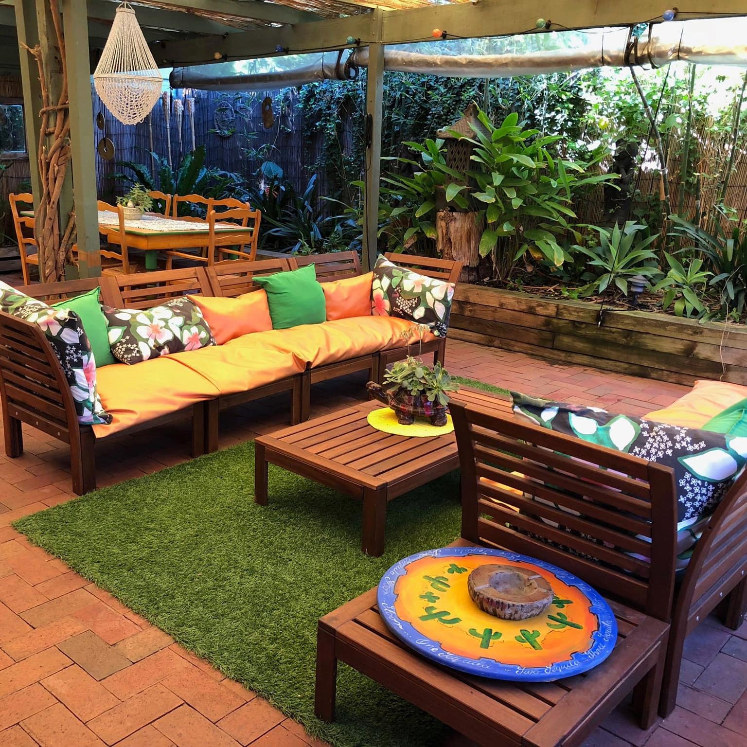 A brick patio space with wooden sofas, colorful cushions, and a wooden dining set, surrounded by lush tropical plants.