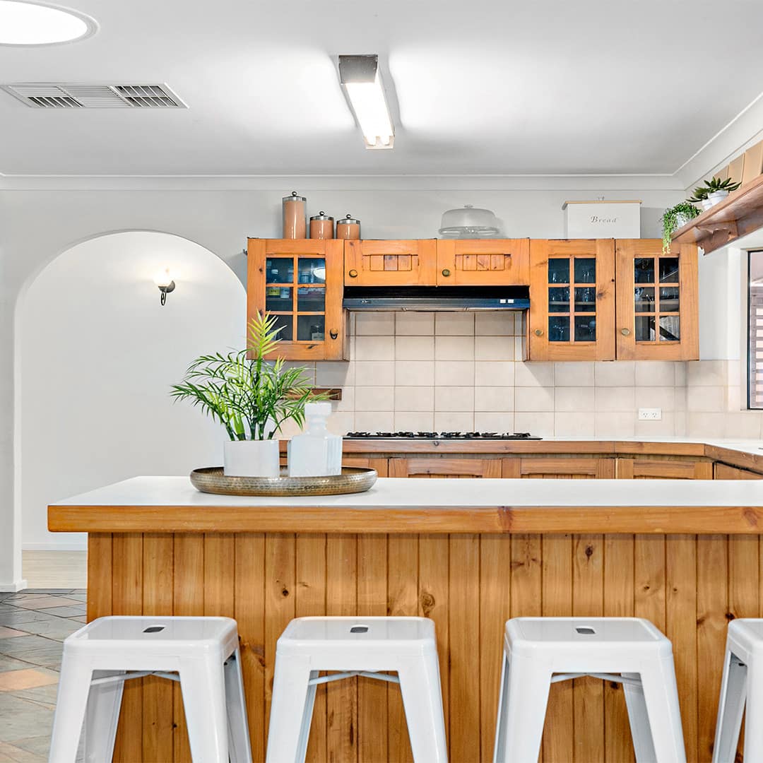 Rustic kitchen with a tan brick accent wall, wood panelled cabinets, white countertops, and white bar stools.