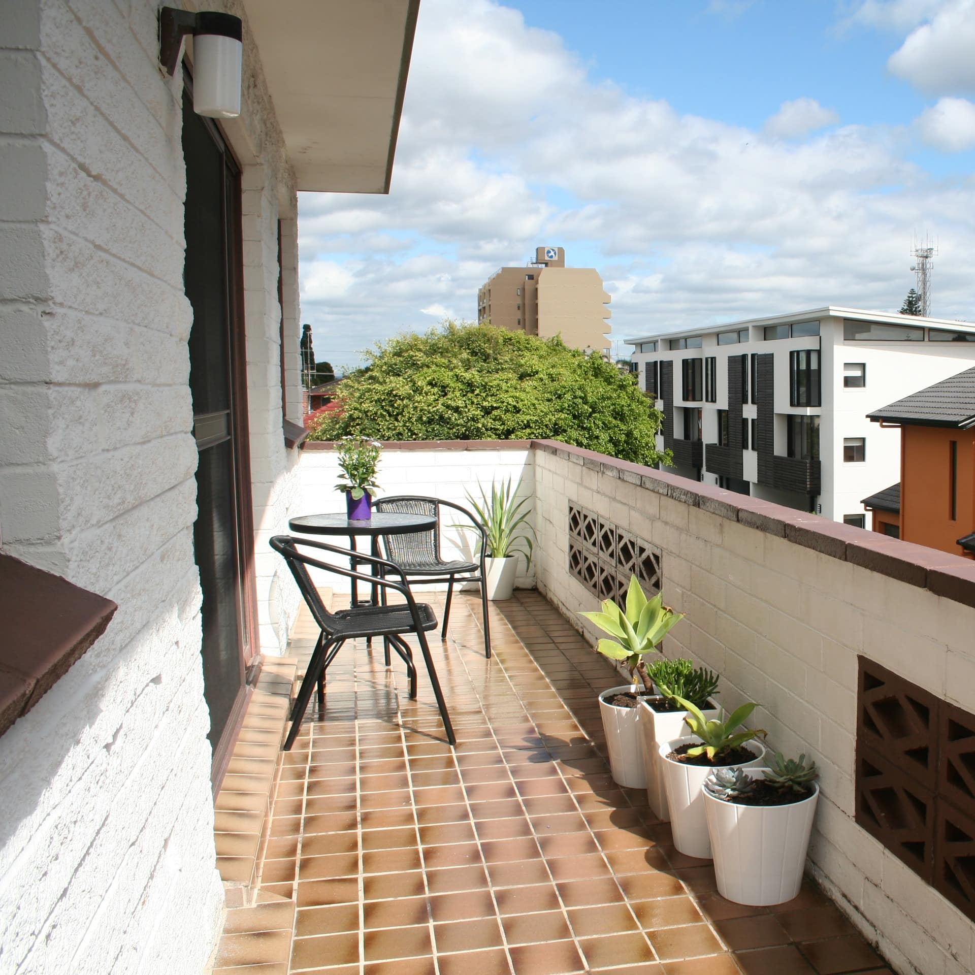A black iron table and two chairs sit on balcony with neighborhood views, brown tile floor, white brick railing.