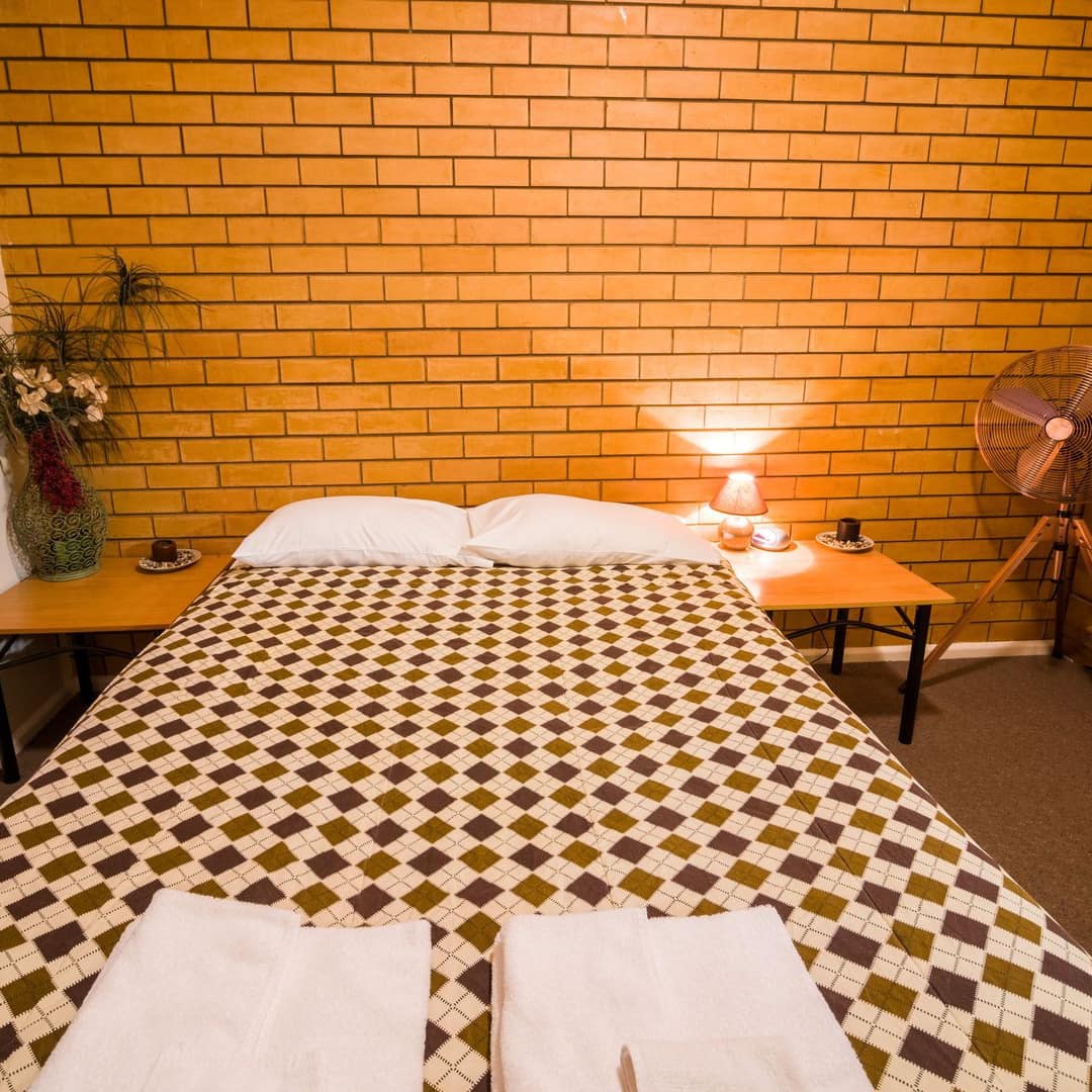 Retro bedroom with orange brick wall, a queen size bed, argyle bedding, matching wooden bedside tables, tall window.