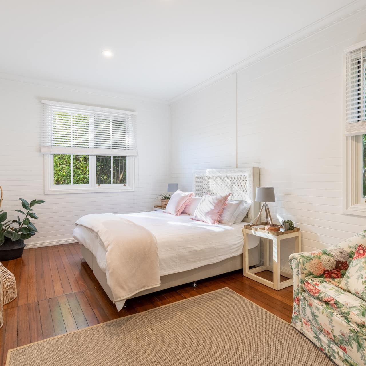 Spacious bedroom with hardwood floors, a king size bed, white and pink bedding, floral armchair, and woven tan rug.