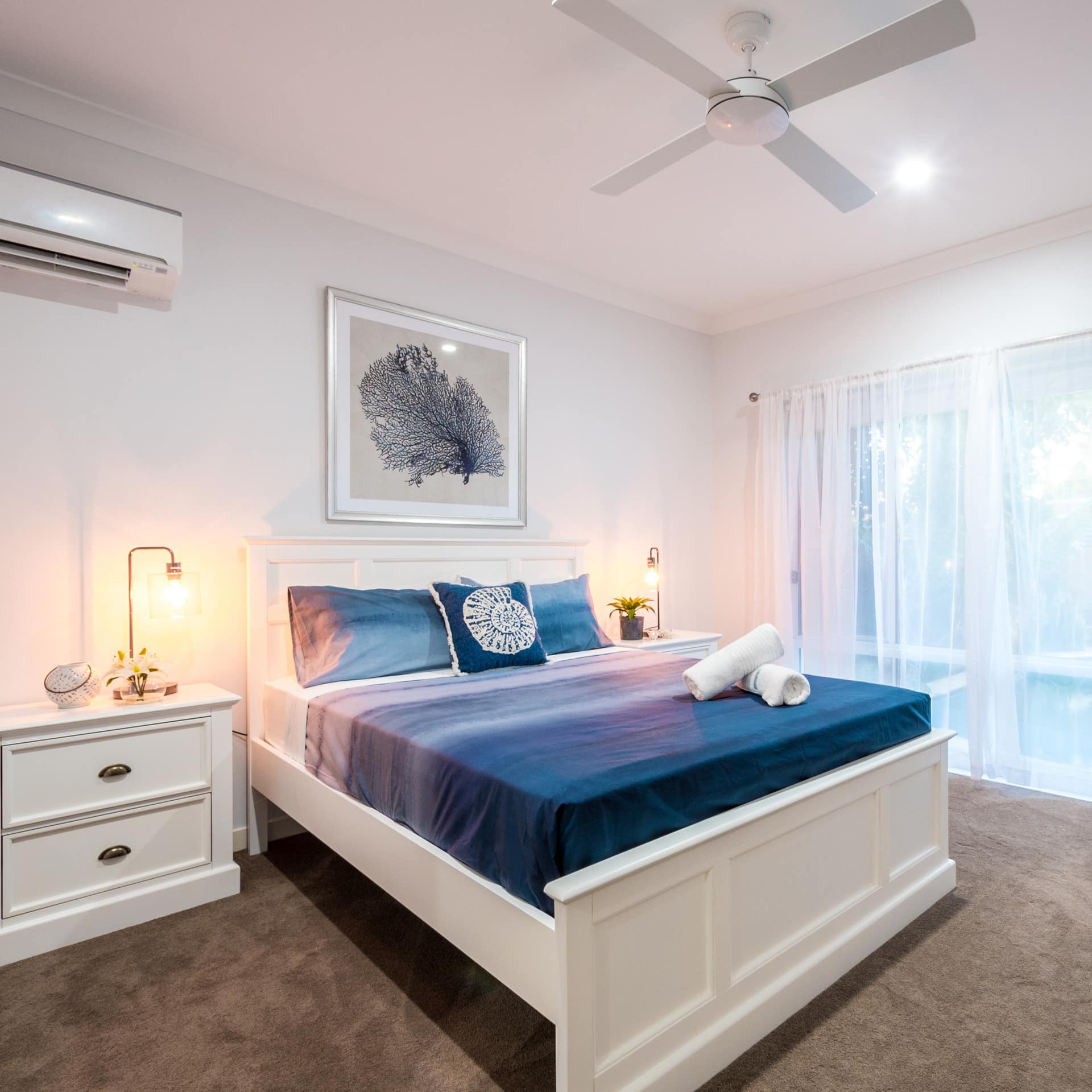 Spacious bedroom with tan carpet, white walls, king size bed, blue bedding, and glass doors to private pool.