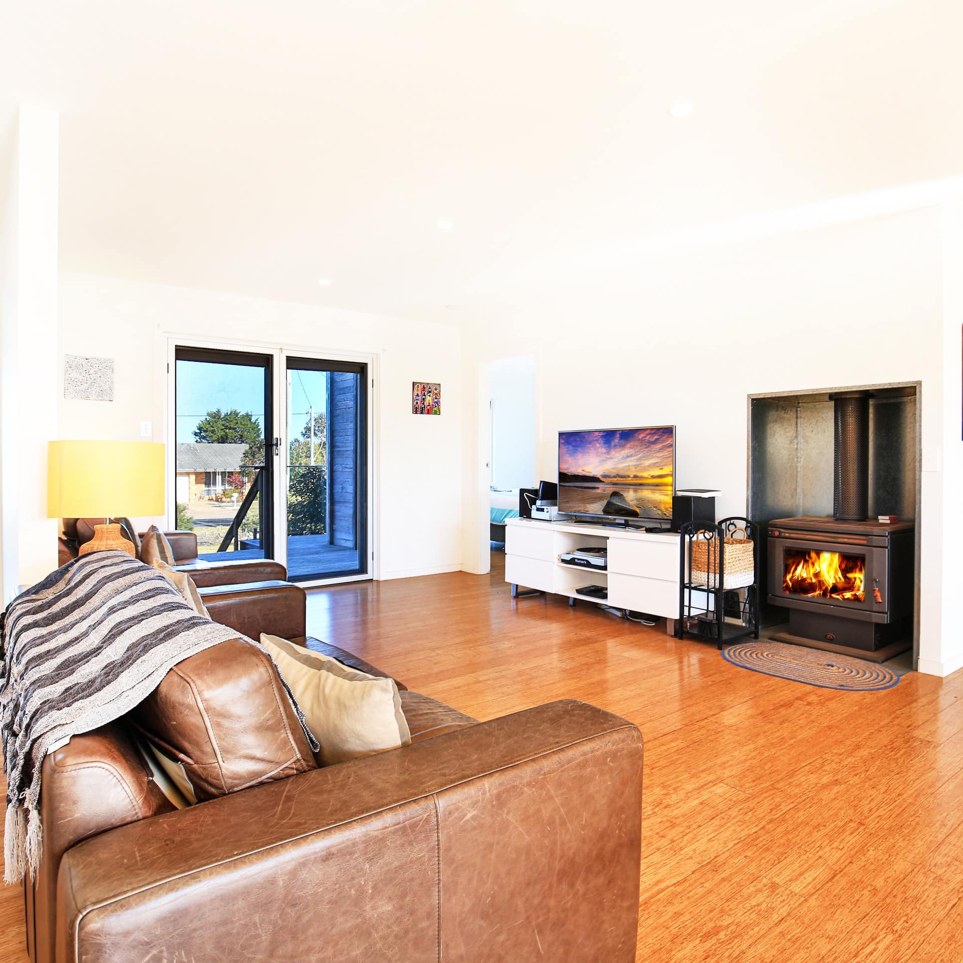 Open living room with hardwood floors, white walls, brown leather sofa, wood-burning stove, and flatscreen TV.