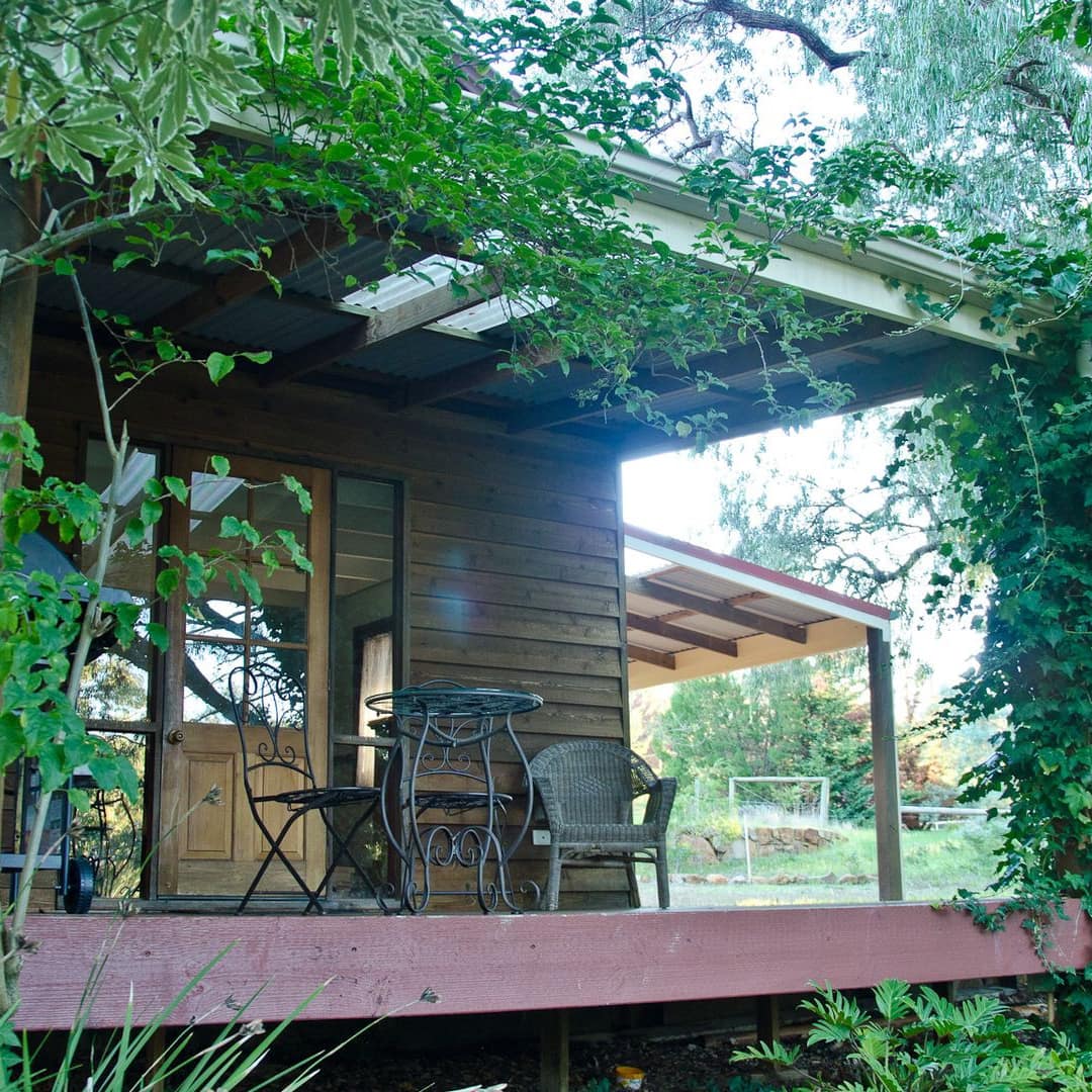A serene cottage in wooded surroundings with a front porch entwined in vines and a small table and chair for drinking coffee