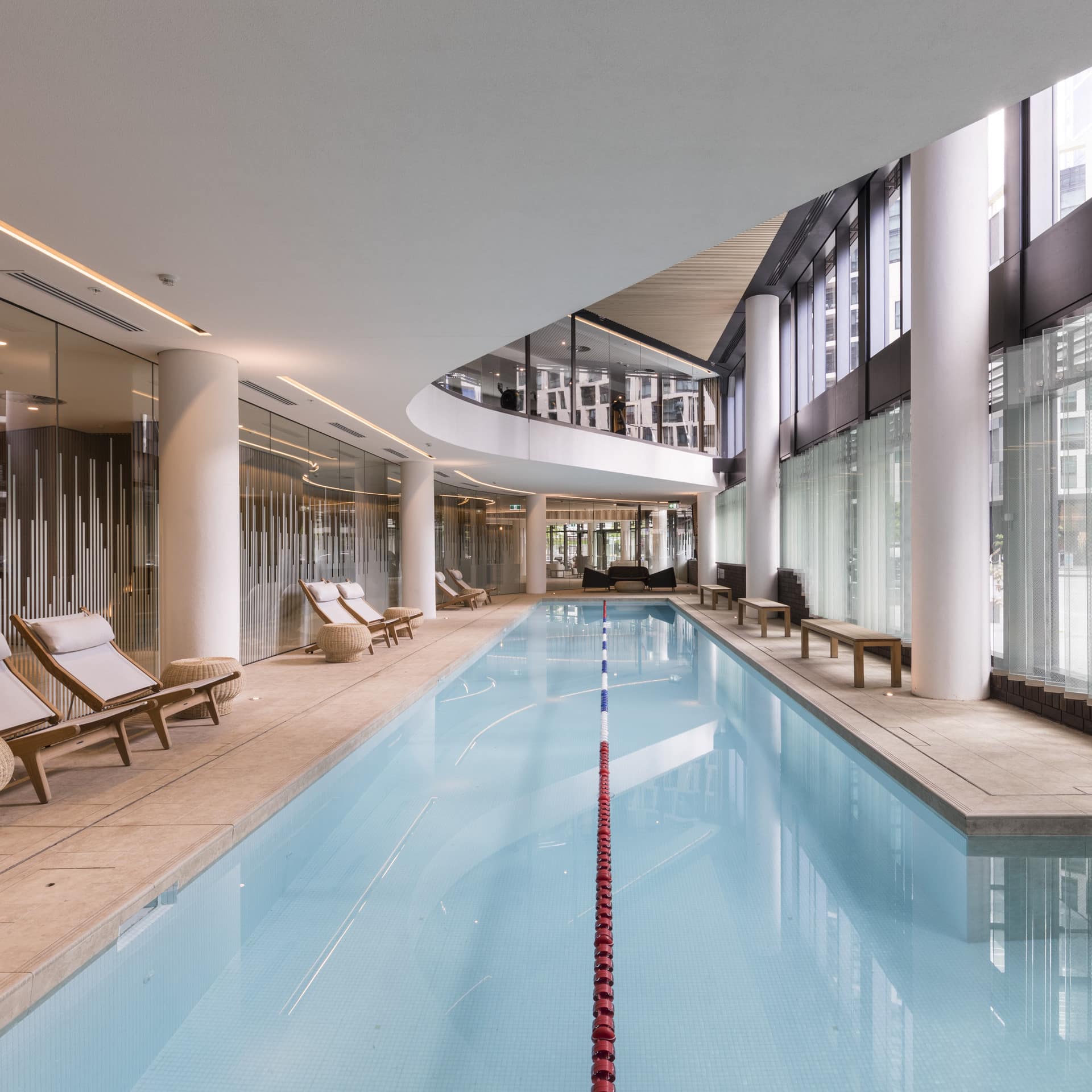 View of long indoor lap pool and beige tile pool deck in a modern building with white canvas lounge chairs.