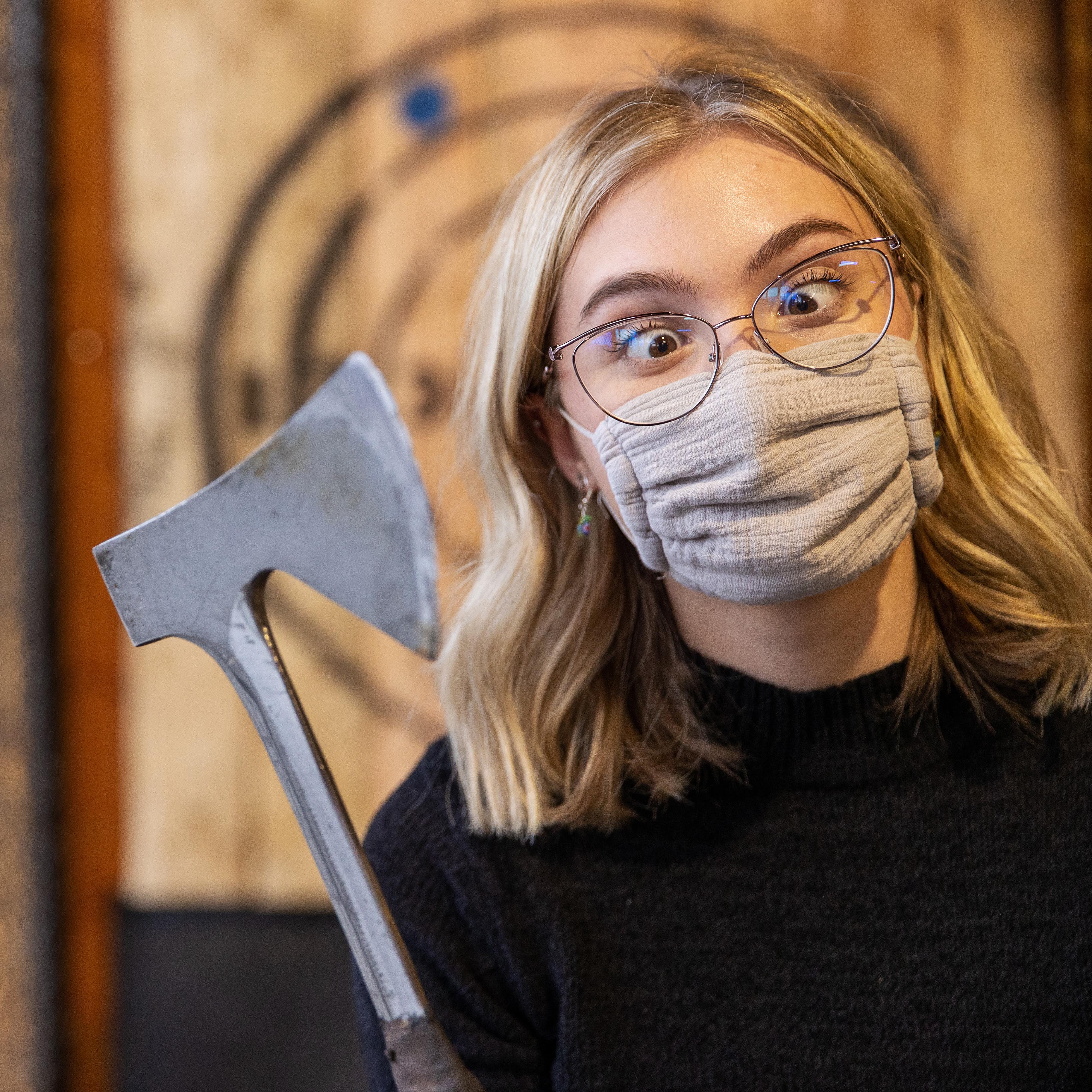 Teenage masked girl wearing glasses with eyes crossed holds axe at an axe-throwing range with target behind her
