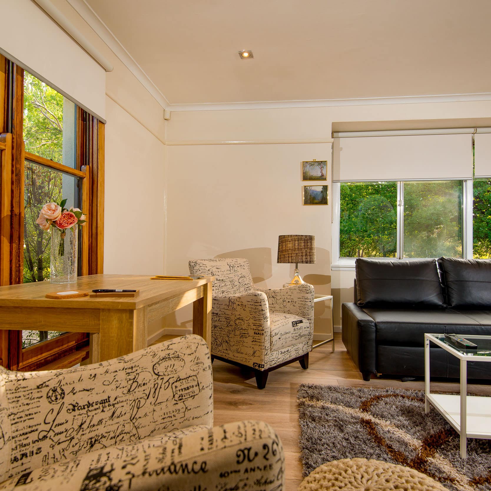 Living room with hardwood floors, leather sofa, white chairs, glass coffee table, and wide windows.