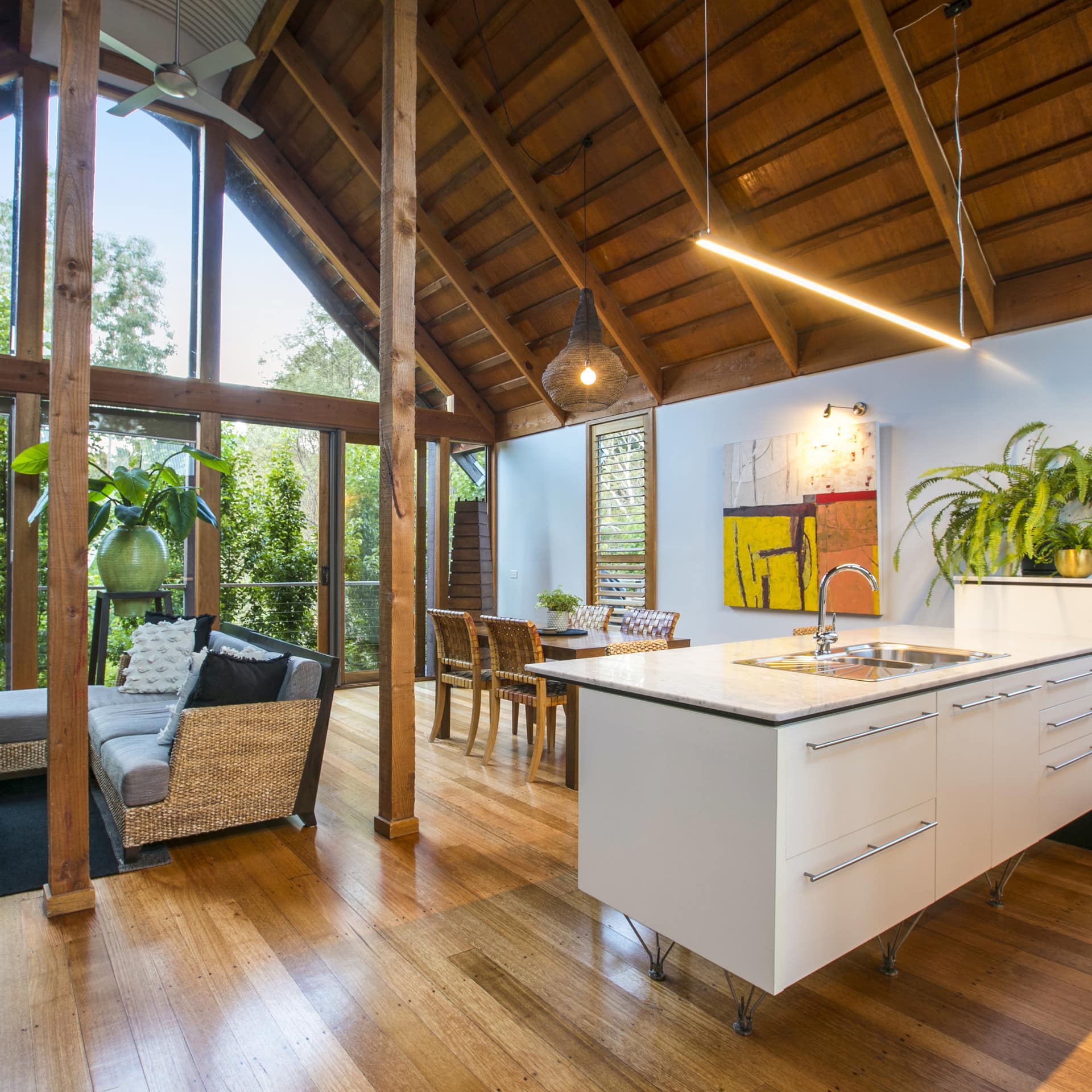 Open living, dining and kitchen area with wood rafters, wood floors, large floor-to-ceiling windows and wicker furniture.