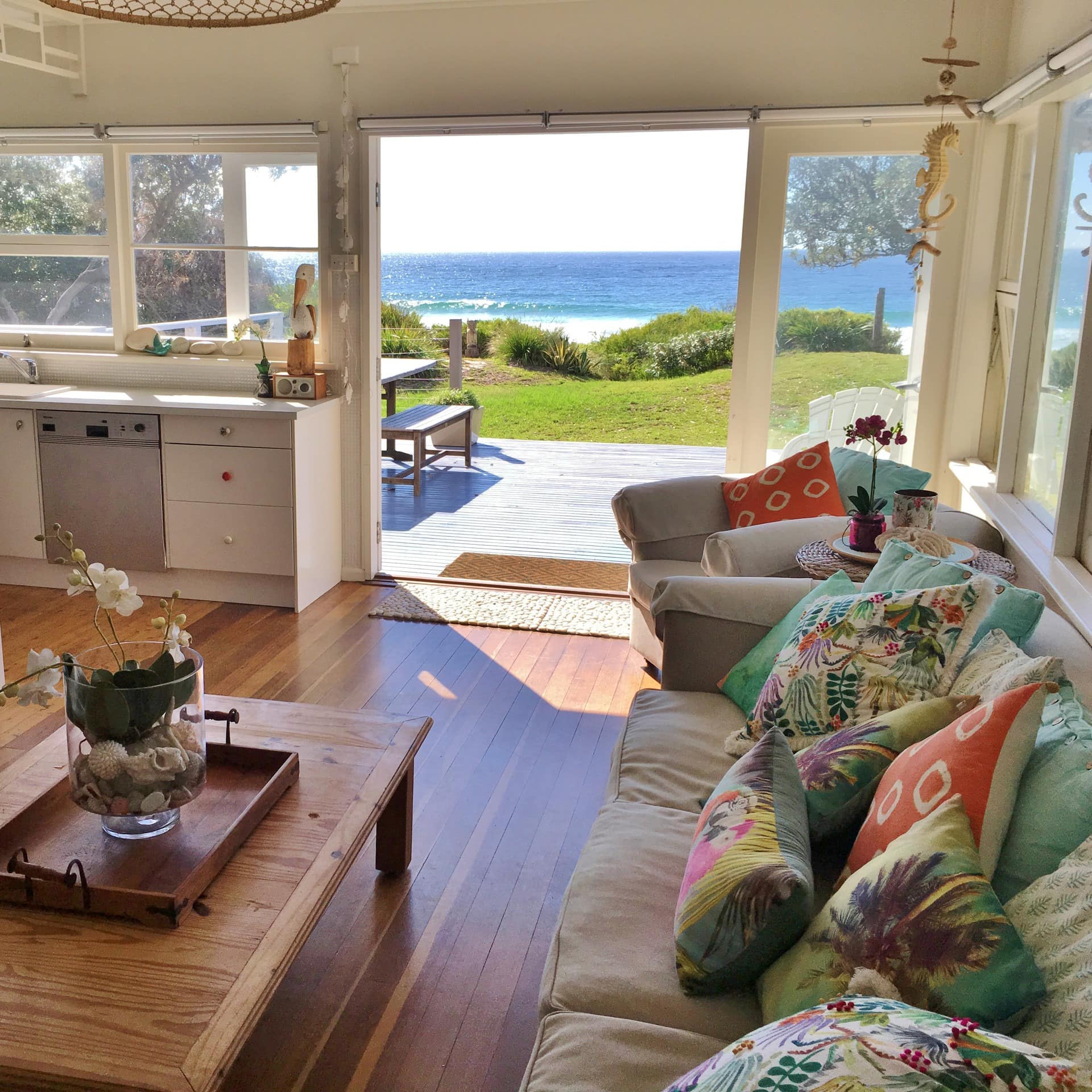  Spacious living room and kitchen, open glass door to patio and ocean views, hardwood floors, colorful decor.