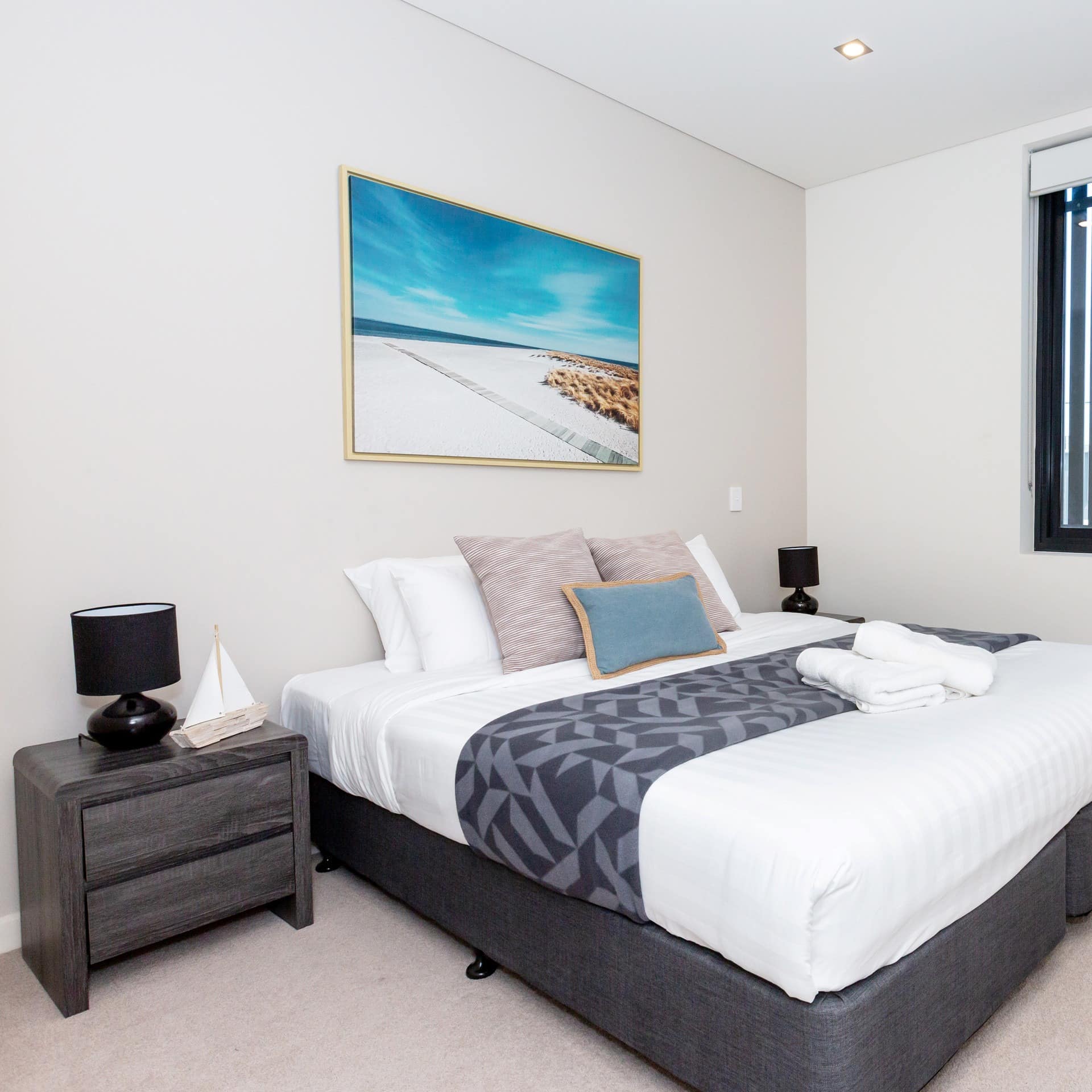 Bedroom with beige carpet and white walls, king-size bed with white and grey bedding, and painting of the beach above the bed.