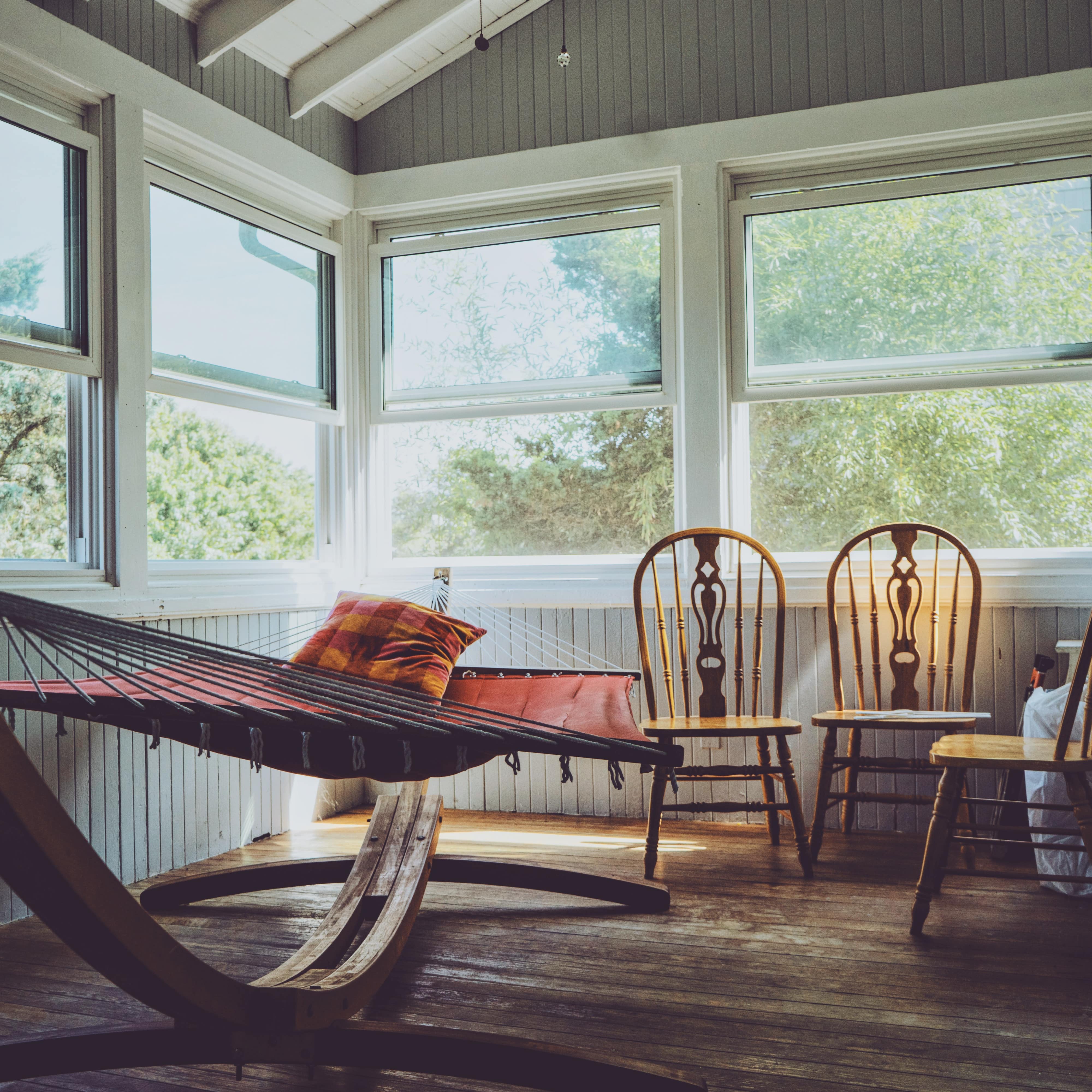 An enclosed porch with windows on two sides, beadboard walls and wood floors with a red hammock on a frame