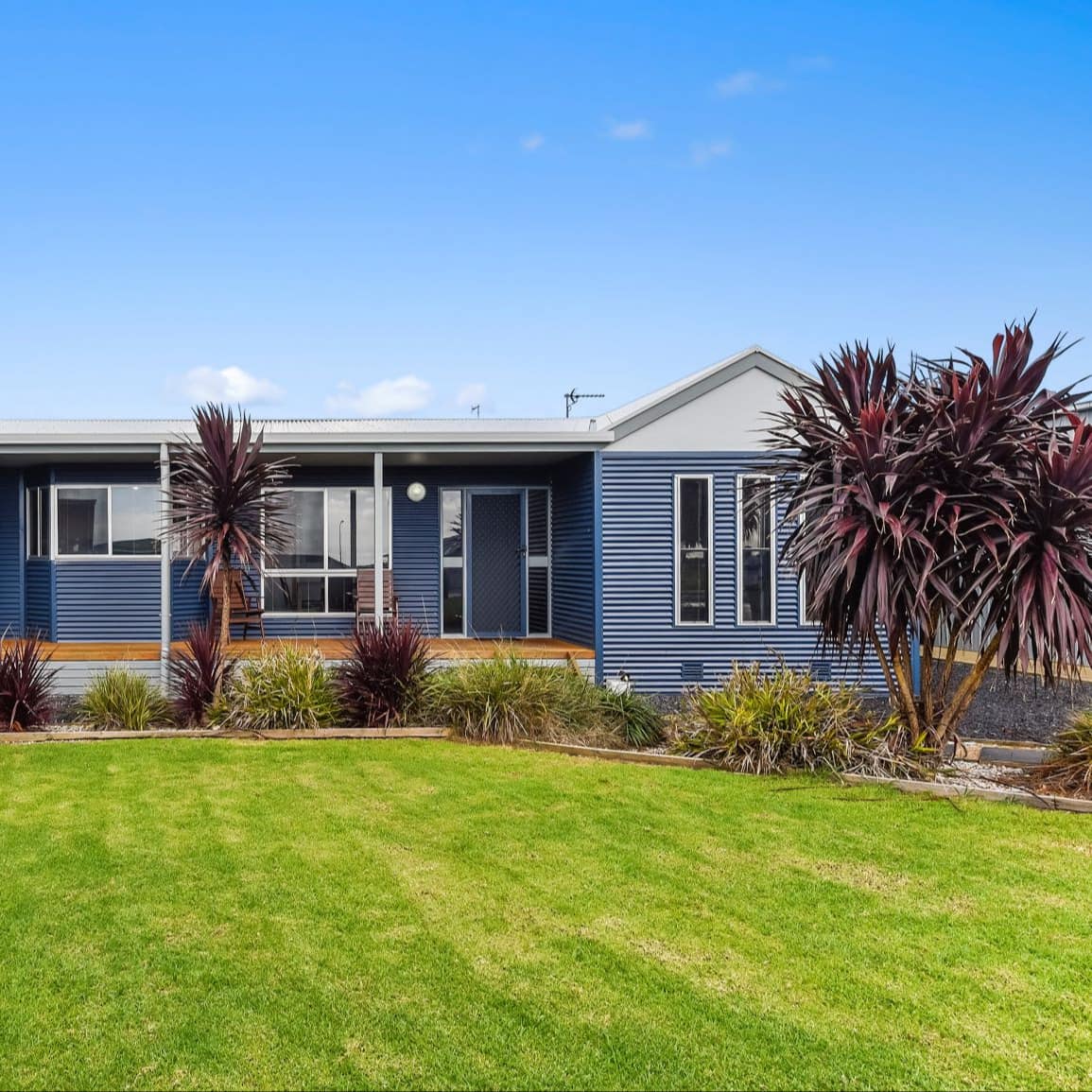 Blue cottage with a white roof, covered front porch, and a manicured lawn with tropical plants.