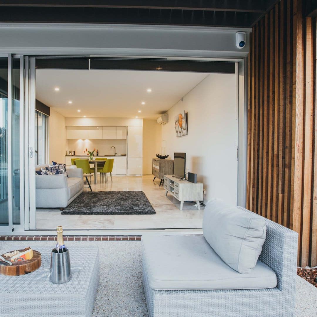 View of a stone patio space with grey wicker furniture and open sliding glass doors to the spacious living room.