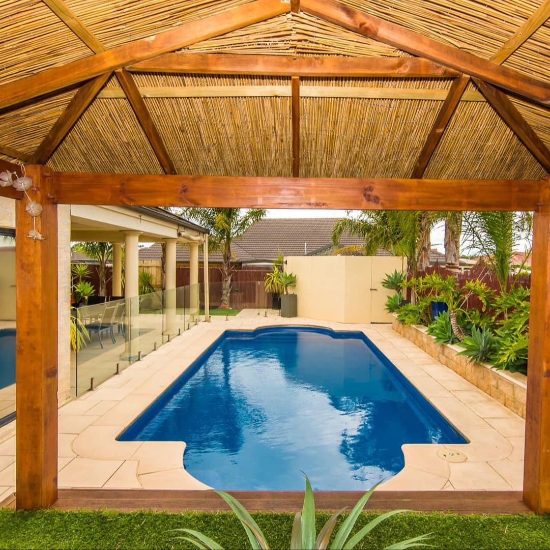 View of private pool with beige tiled pool deck, straw cabana, raised plant beds with tropical plants.