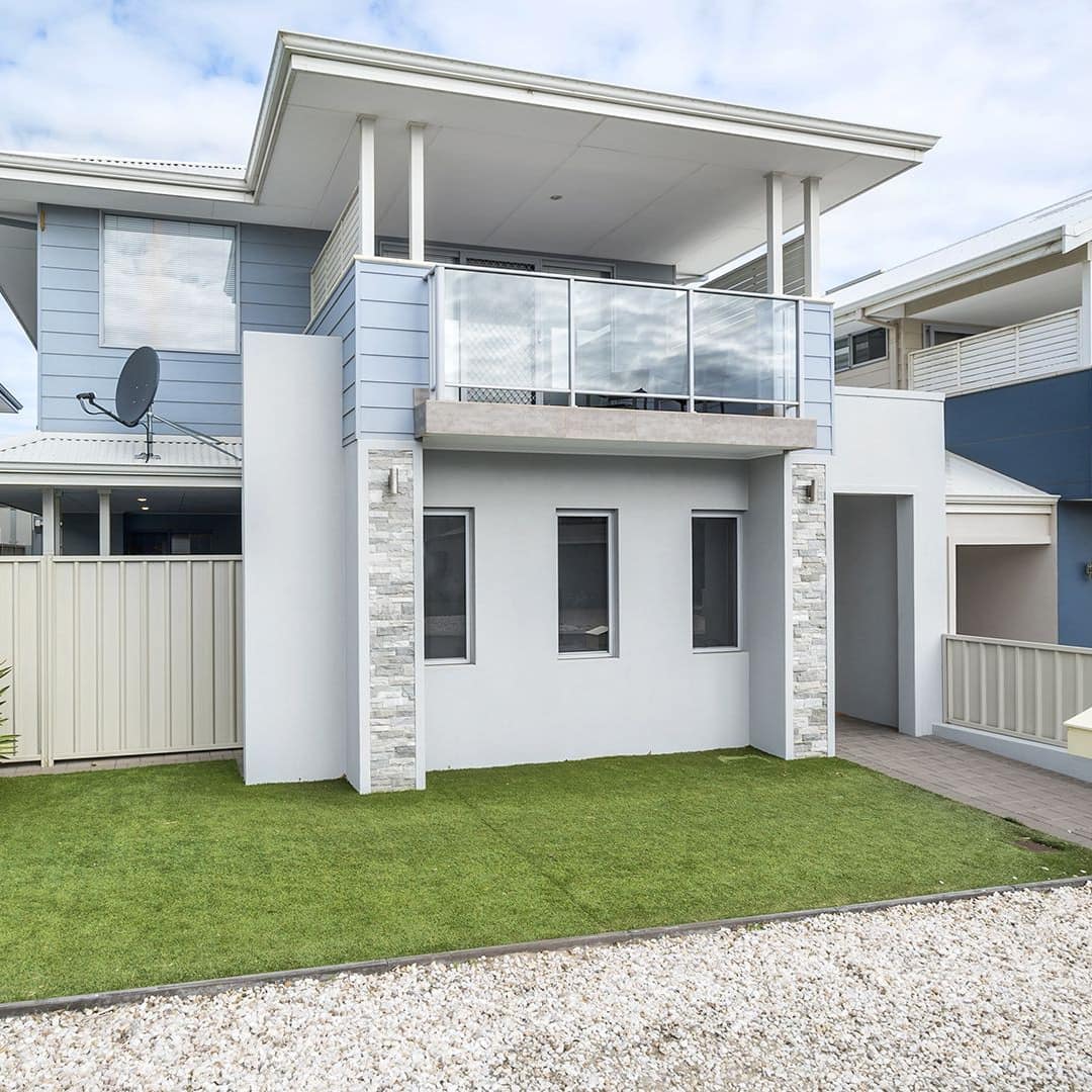 Exterior of a modern two-story home with blue and white siding, landscaped lawn with tropical plants and gravel driveway.