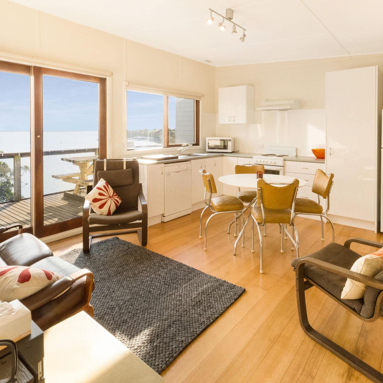 Open living room and kitchen, dining area, hardwood floors, brown canvas furniture, attached verandah with ocean views.