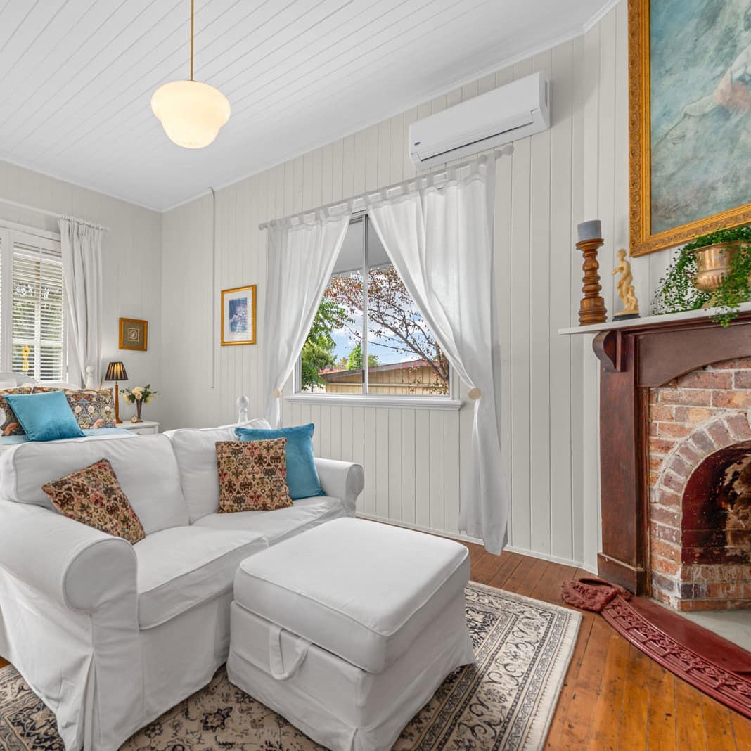 Farmhouse-style bedroom with hardwood floors, white panelled walls, king size bed, white sofa, brick fireplace.
