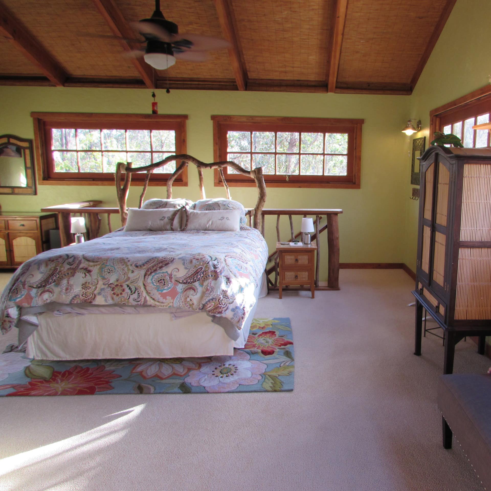 Spacious bedroom with beige carpet, light green walls, a king size bed with natural wood bed frame, and tropical decor.