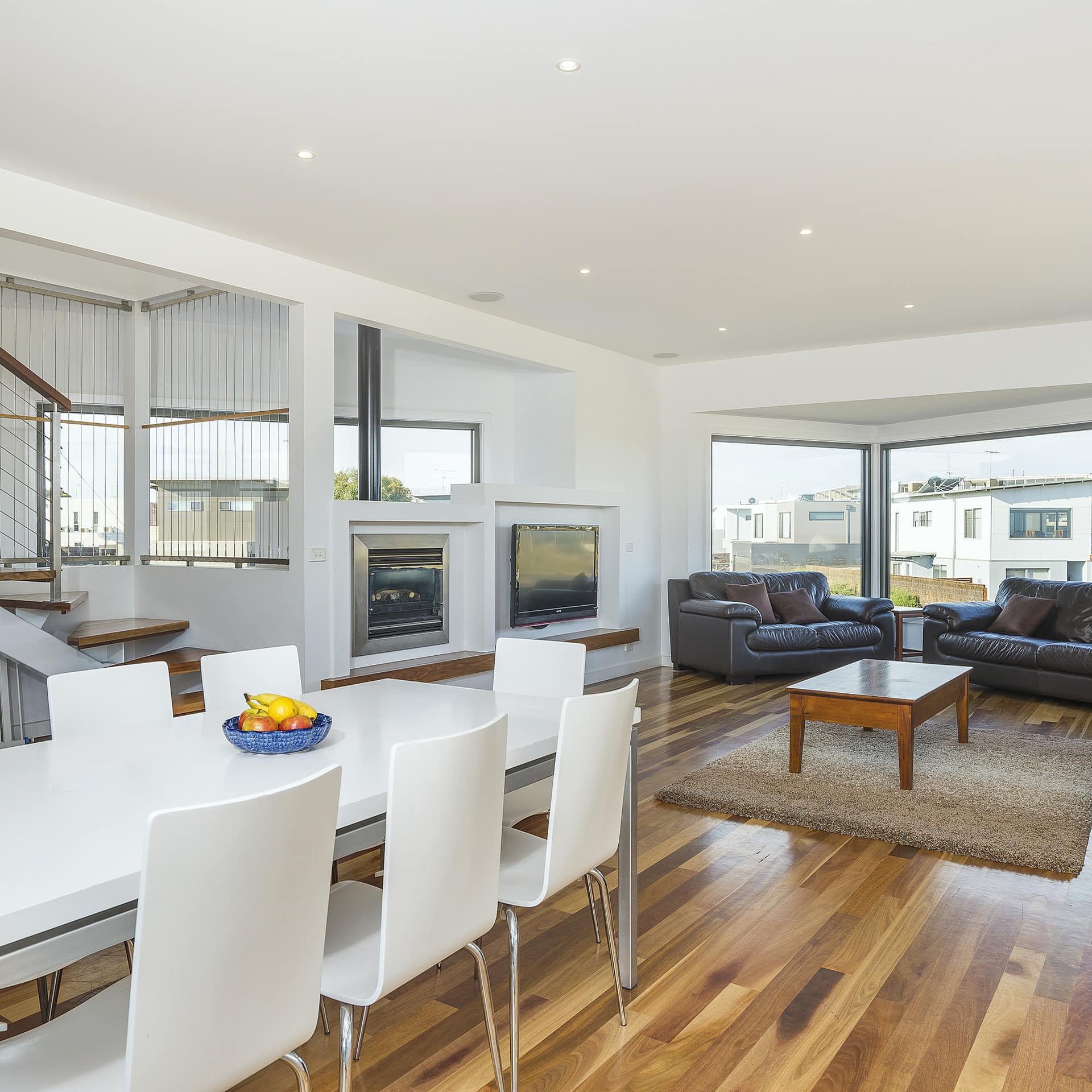 Modern living and dining room with hardwood floors, black leather sofas, white dining set, fireplace, and flatscreen TV.