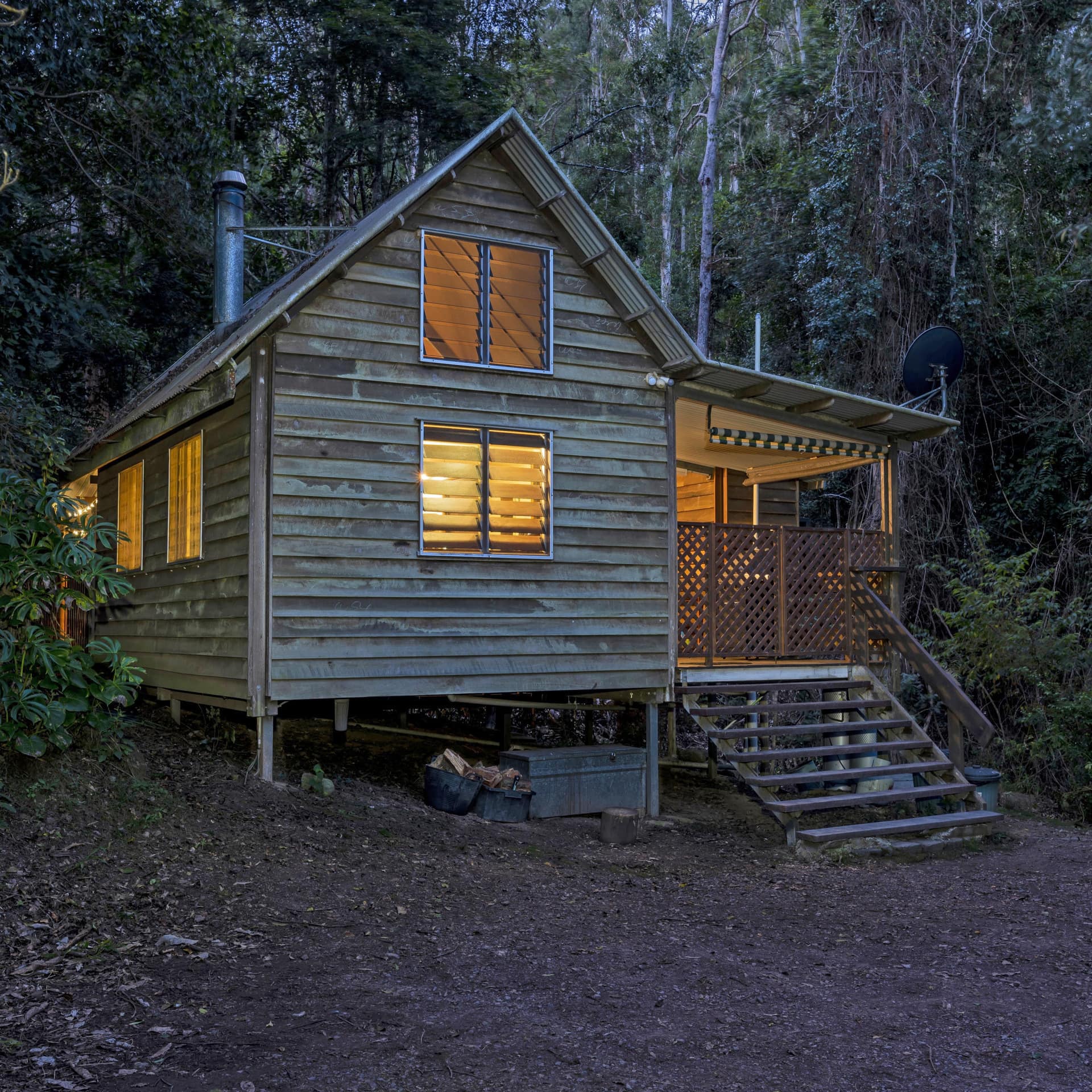 An evening view of a quaint wood cabin with a covered deck, surrounded by thick forest.