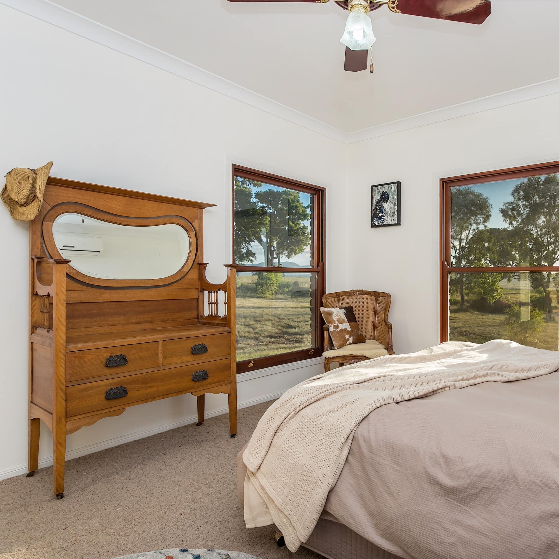 Rustic bedroom with a king size bed, beige bedding, vanity, a wicker chair with a cowhide pillow, windows with rural views.