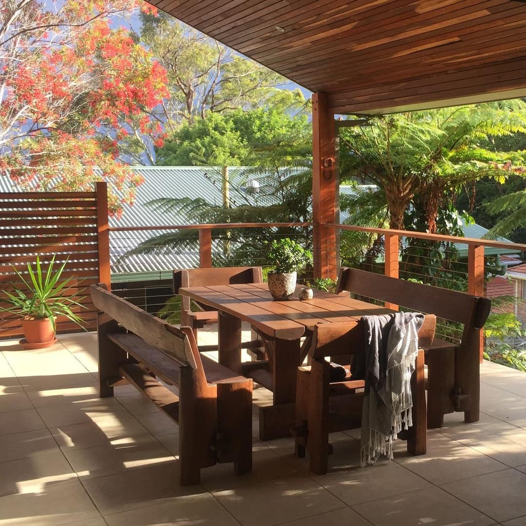 Cosy patio with wooden table, benches, railing and sloped roof surrounded by green plants and views of trees and blue sky