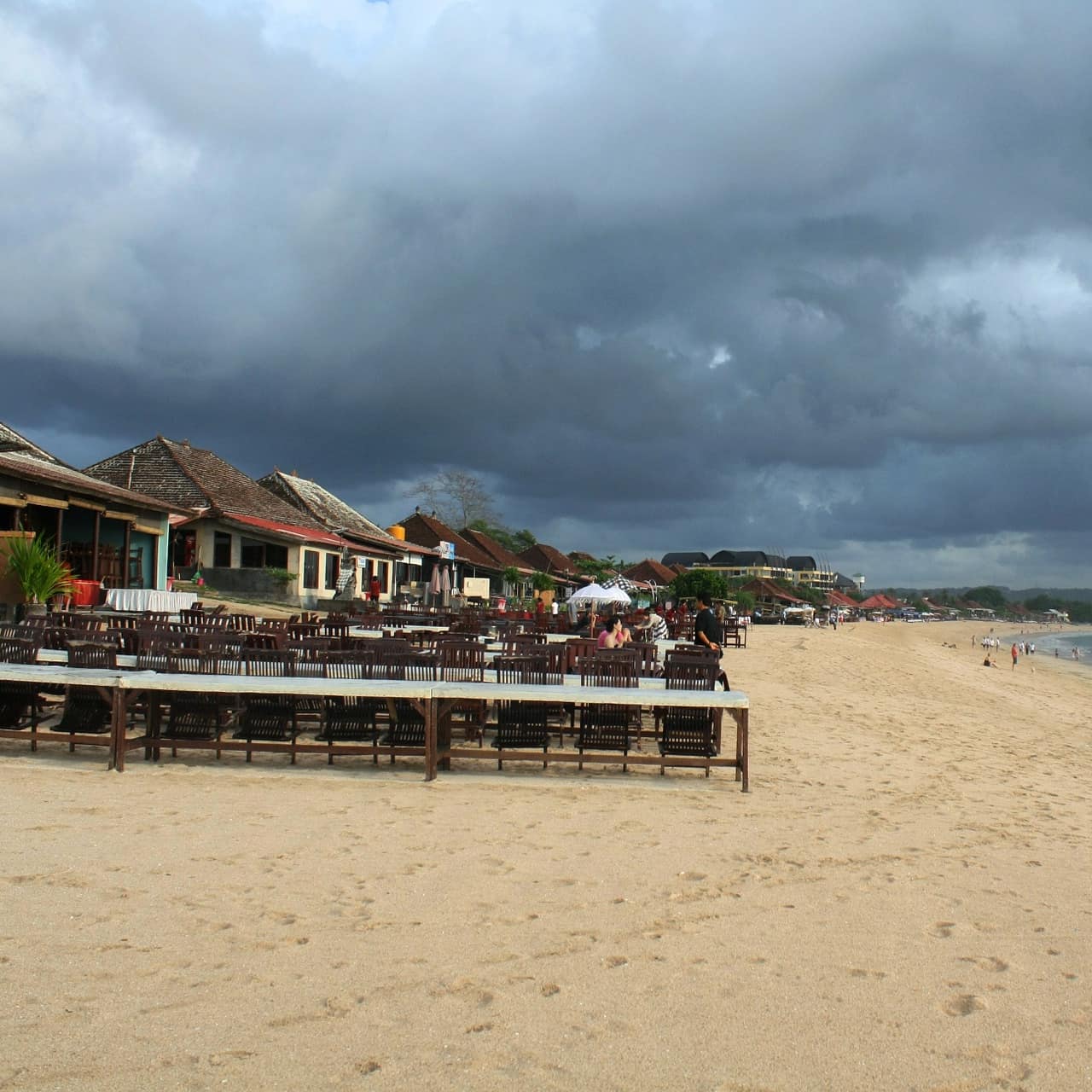 Bali beachfront villas in a row with tables for eating outdoors on the beach with people enjoying swimming and stormy skies