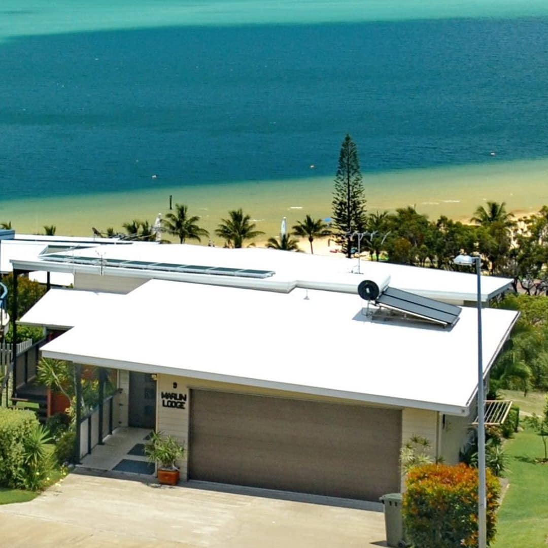 Exterior view of Marlin Lodge with beige siding, white roof, beige cement driveway, garage, and the ocean behind the home.