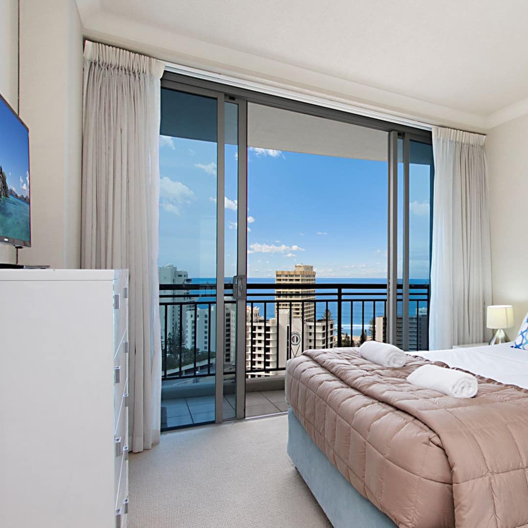 Bedroom with beige carpet, king size bed with blue suede frame, white and tan bedding, flatscreen TV, attached balcony.