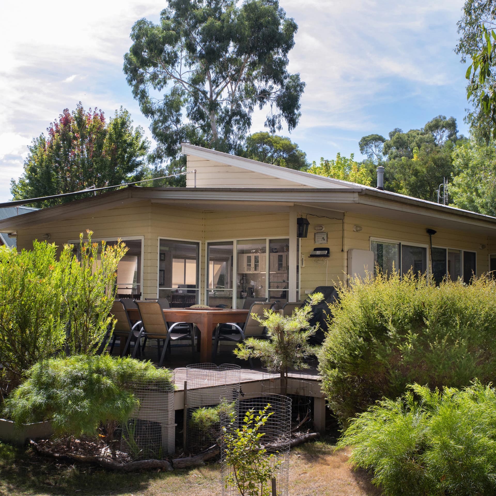 A yellow cottage with a deck, wooden outdoor dining set, and lush yard.