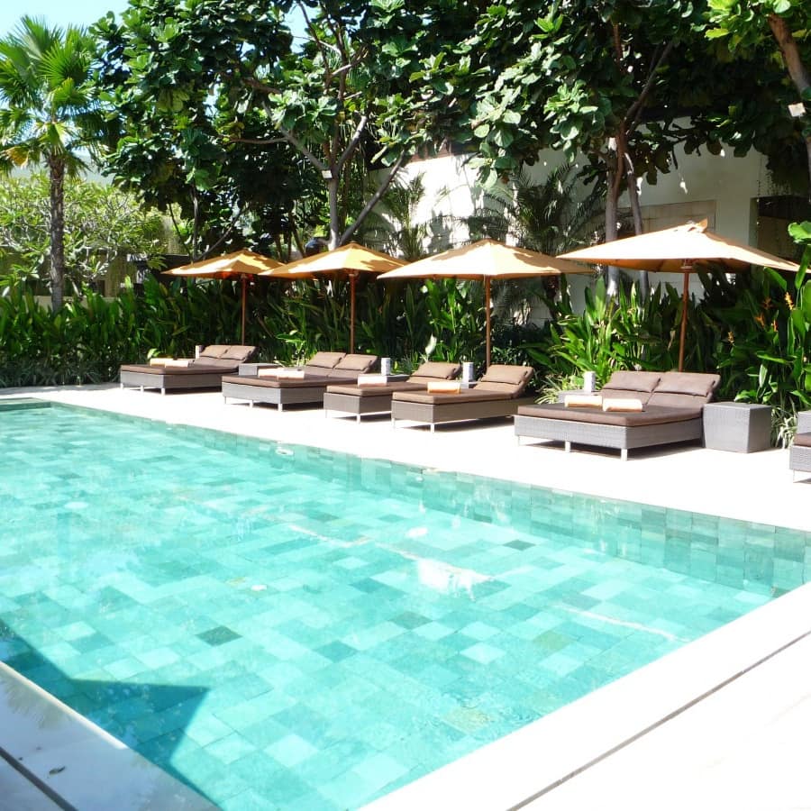 A bright aqua pool with patchwork tiles, brown lounge chairs and shade umbrellas with tropical plants and a white villa