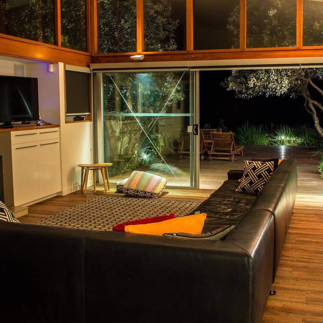 Open living room with black leather sectional, flatscreen TV, and floor to ceiling windows, glass doors to backyard.