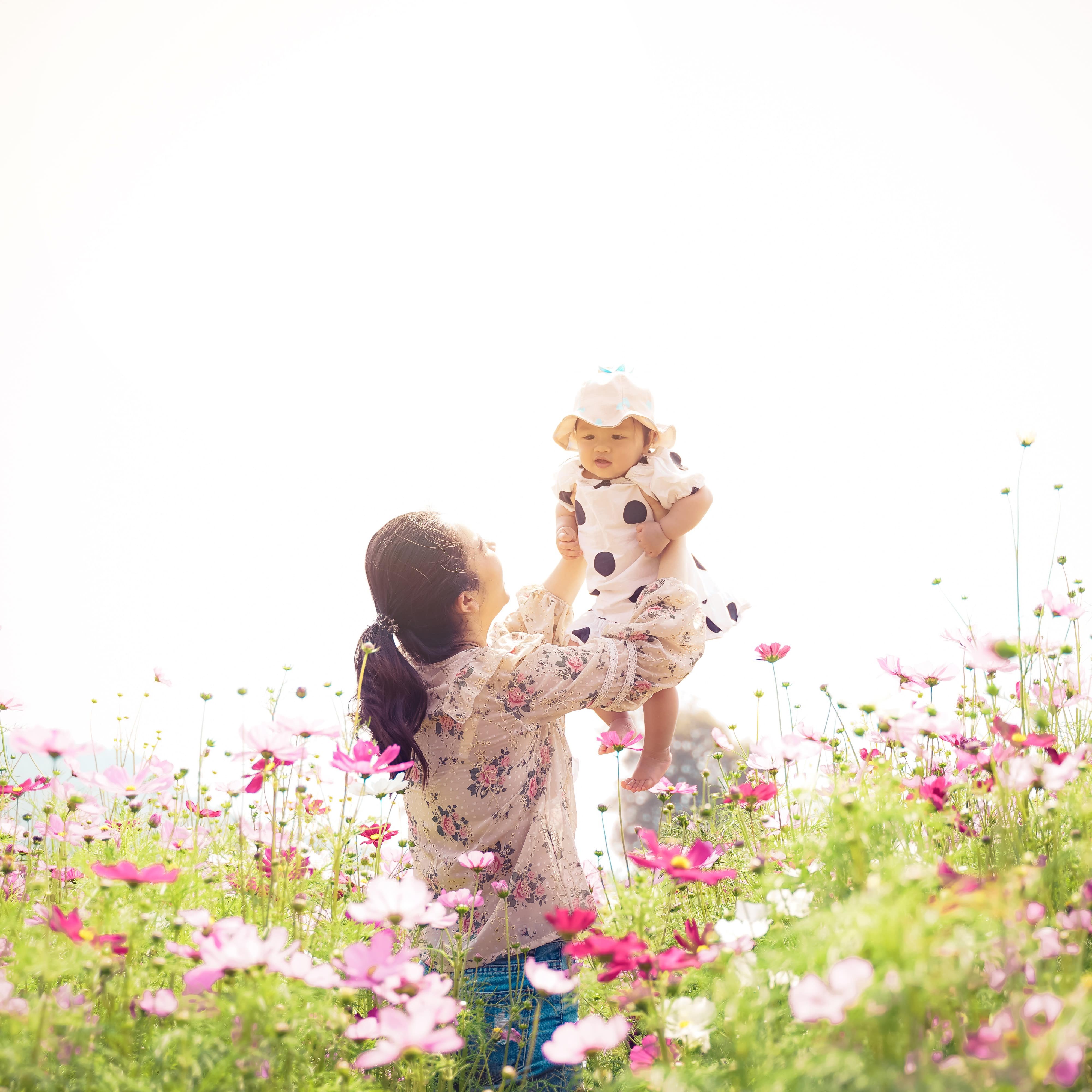 A mother holding her infant daughter in the air in garden of spring wildflowers of pink blossoms at sunset time