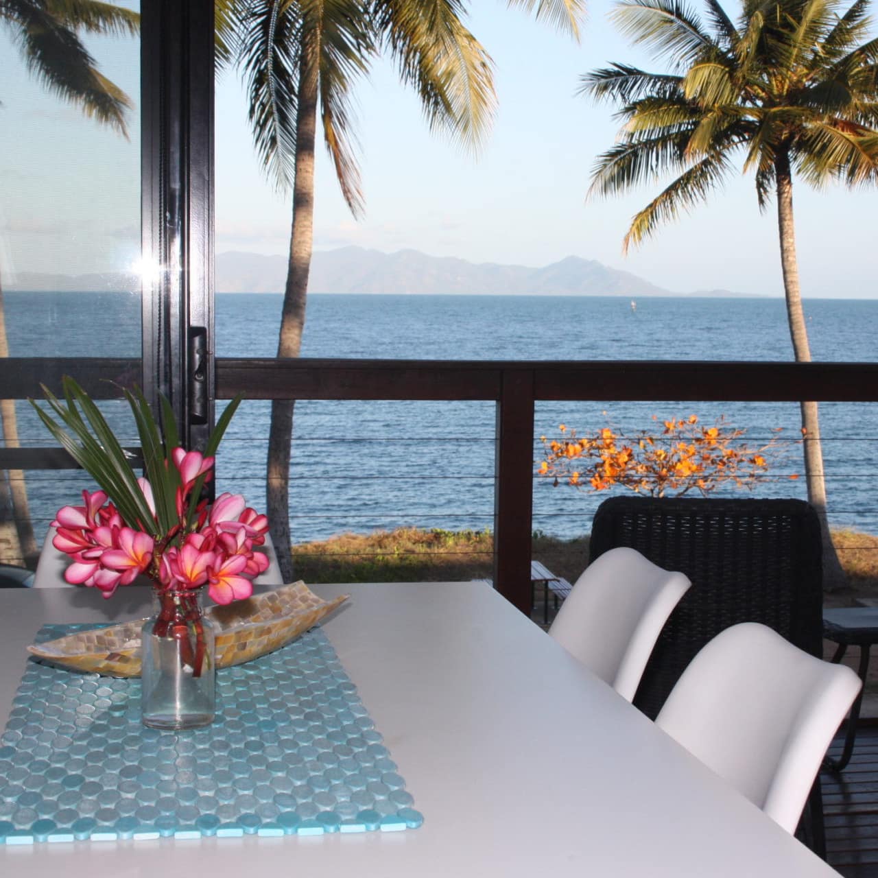 View of the ocean and palm trees through the open sliding glass door of a dining room with a white dining set.