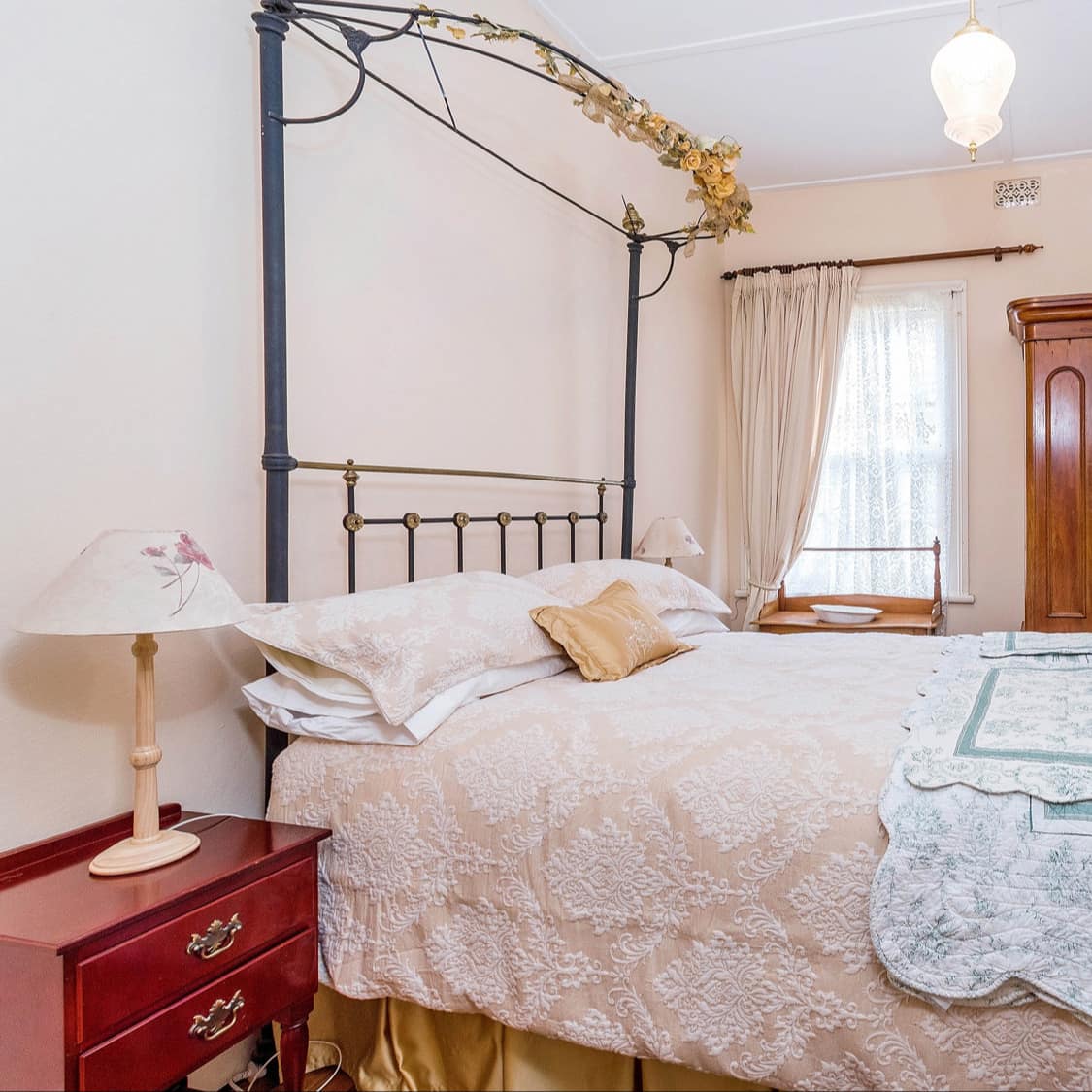 Master bedroom with a wrought iron bed, peach linens, an antique wardrobe, a curtained window, and a cherry bedside table.