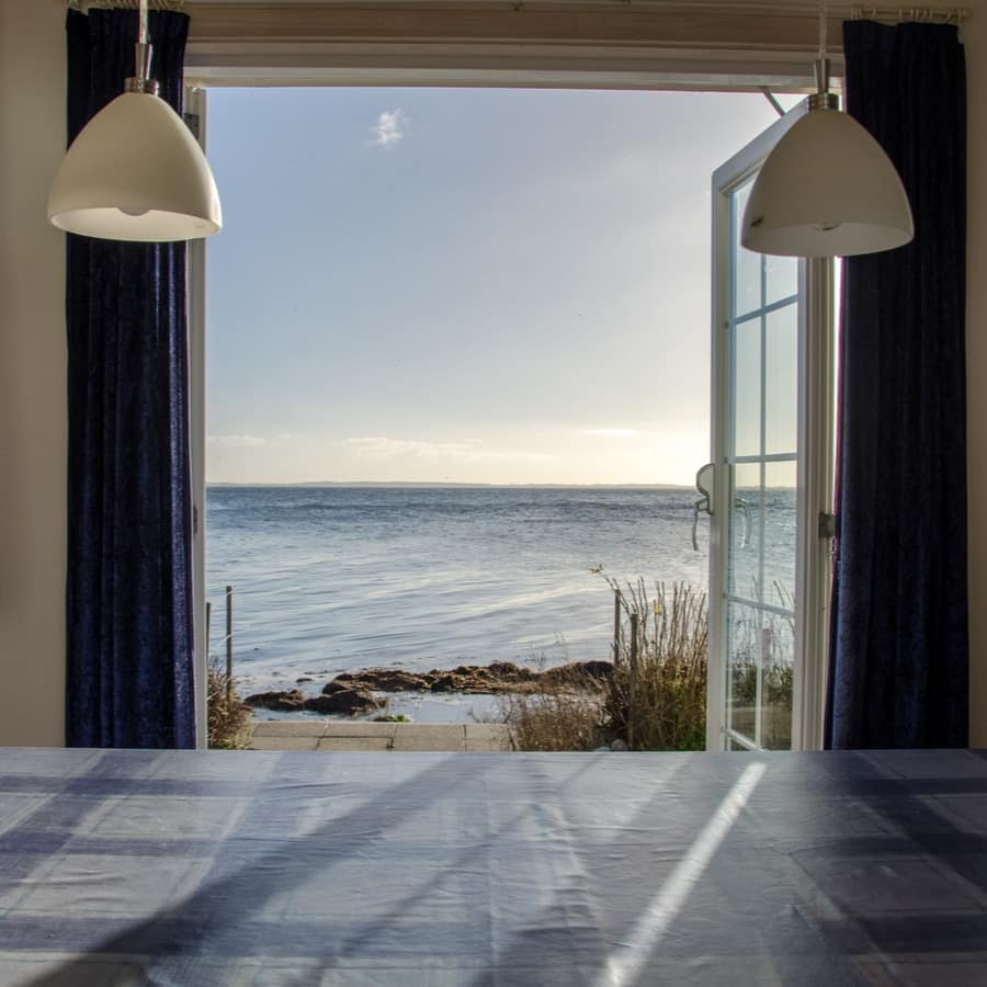 Dining table with blue tablecloth and curtains of beachfront apartment with open doors to patio and ocean