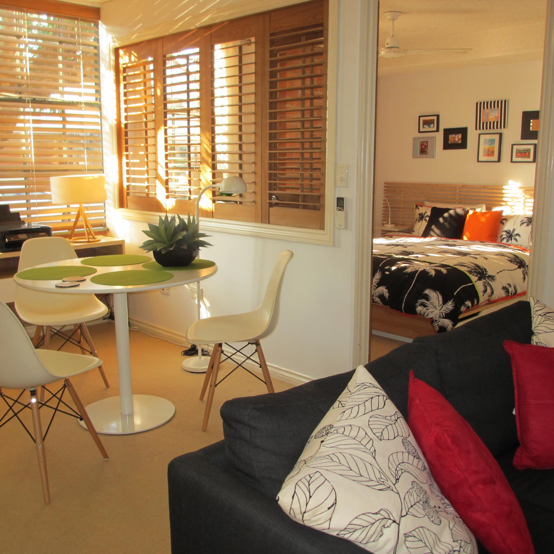 Sunny dining and living room and open door to bedroom, tropical and modern decor, wide windows with wood blinds.