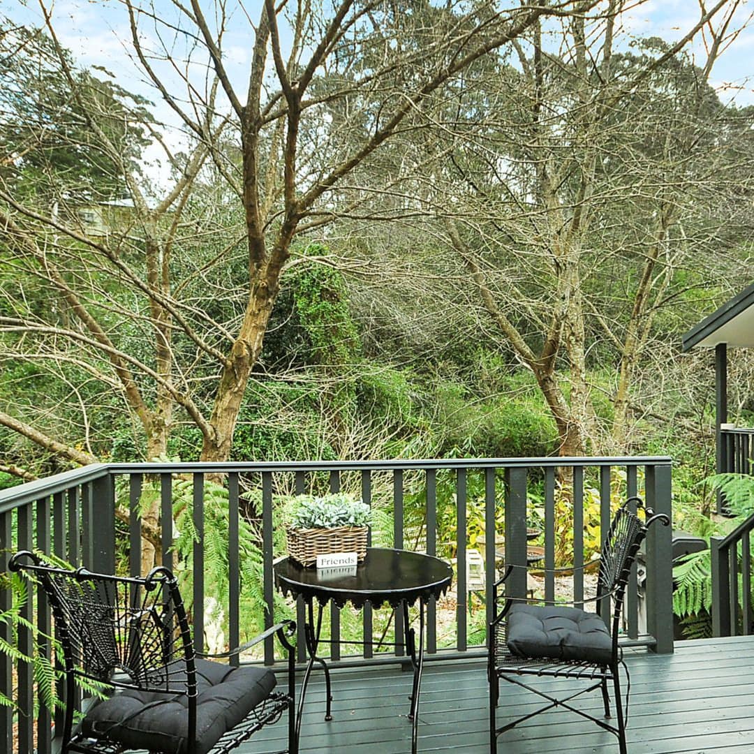 View of the deck of a white cottage with a black metal dining set, and surrounded by thick forest.