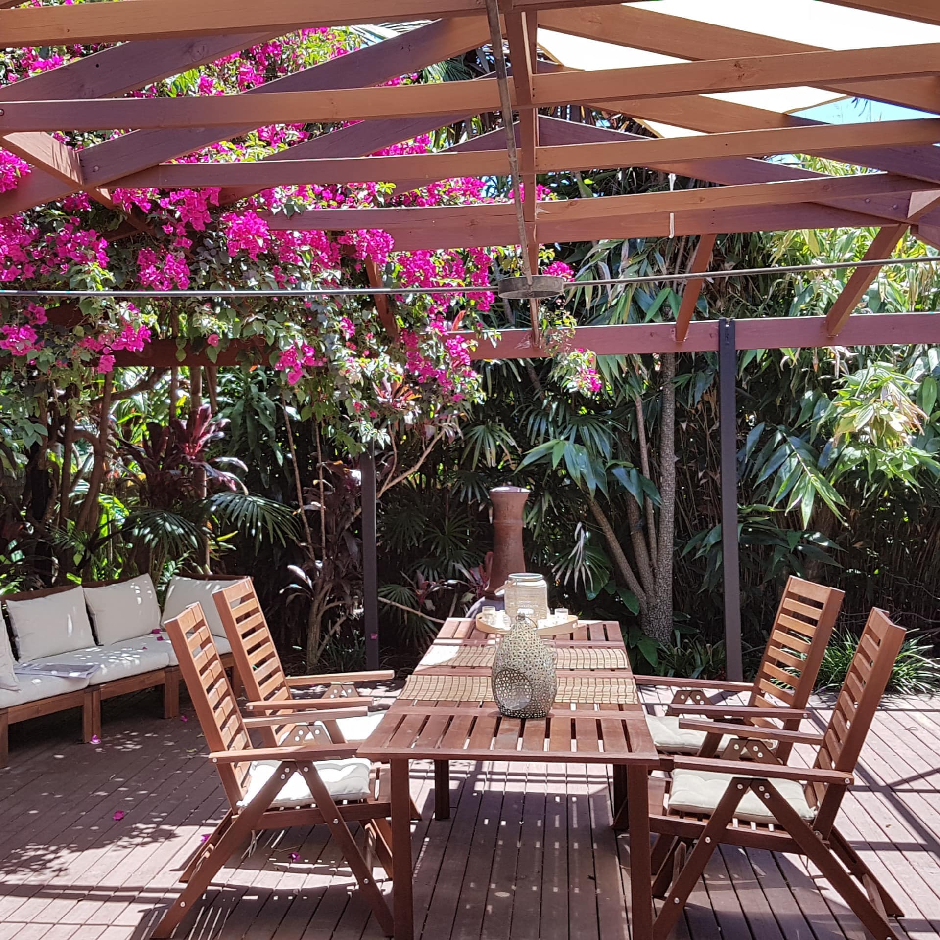 Wooden pergola and deck space surrounded by lush, tropical vegetation with wood dining set and white canvas sofa.