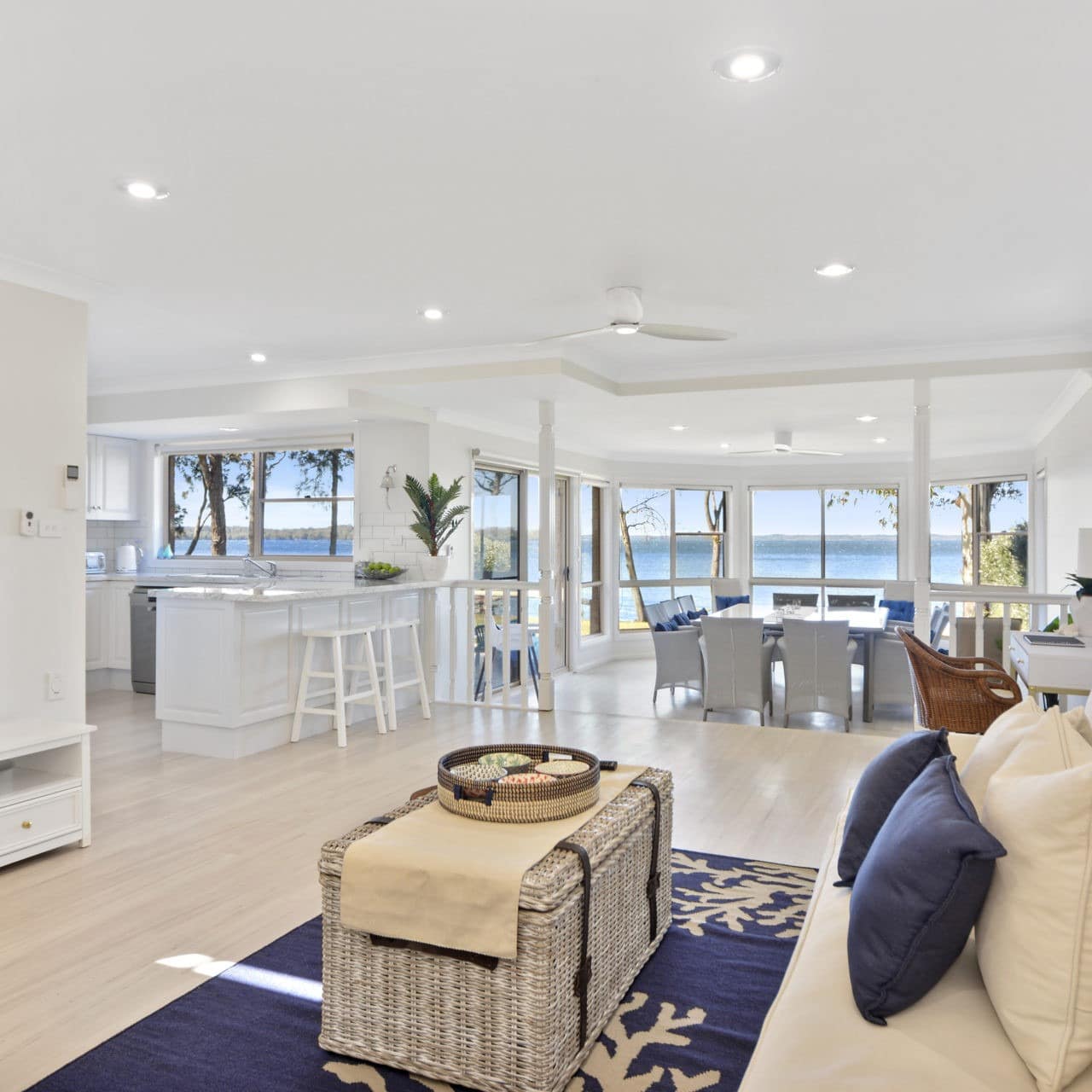 Hamptons-inspired living and dining rooms with coastal furnishings, wicker coffee table, blue accents and waterfront view. 