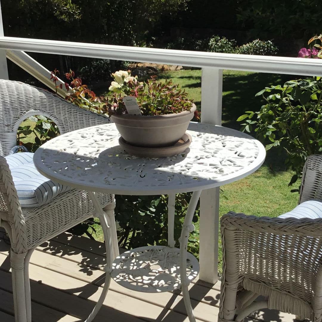 A white, wrought iron table with two wicker chairs sit on the covered deck of the cottage, overlooking the private garden.