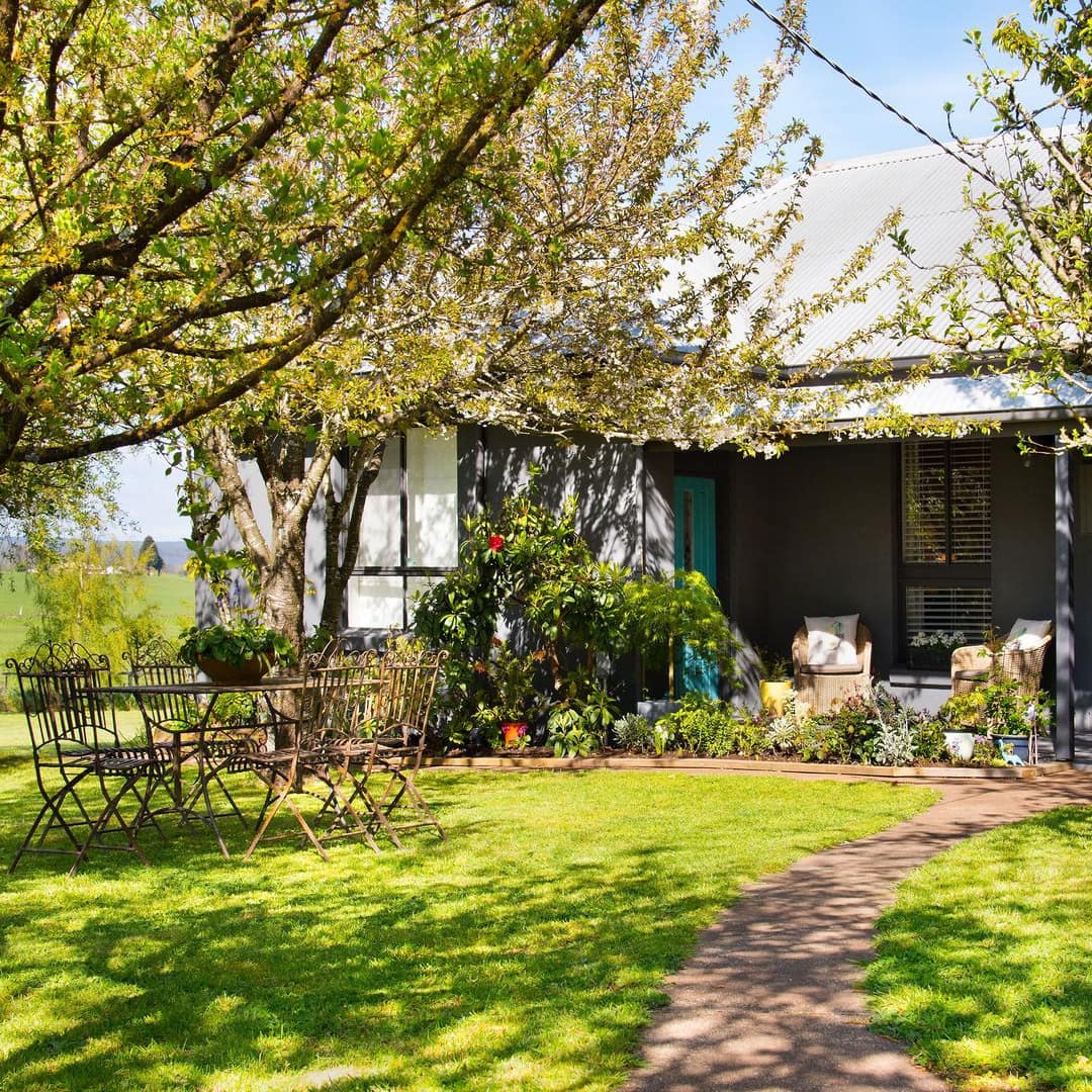 Sunlit front lawn of cottage with covered front porch, black metal dining set on landscaped lawn.