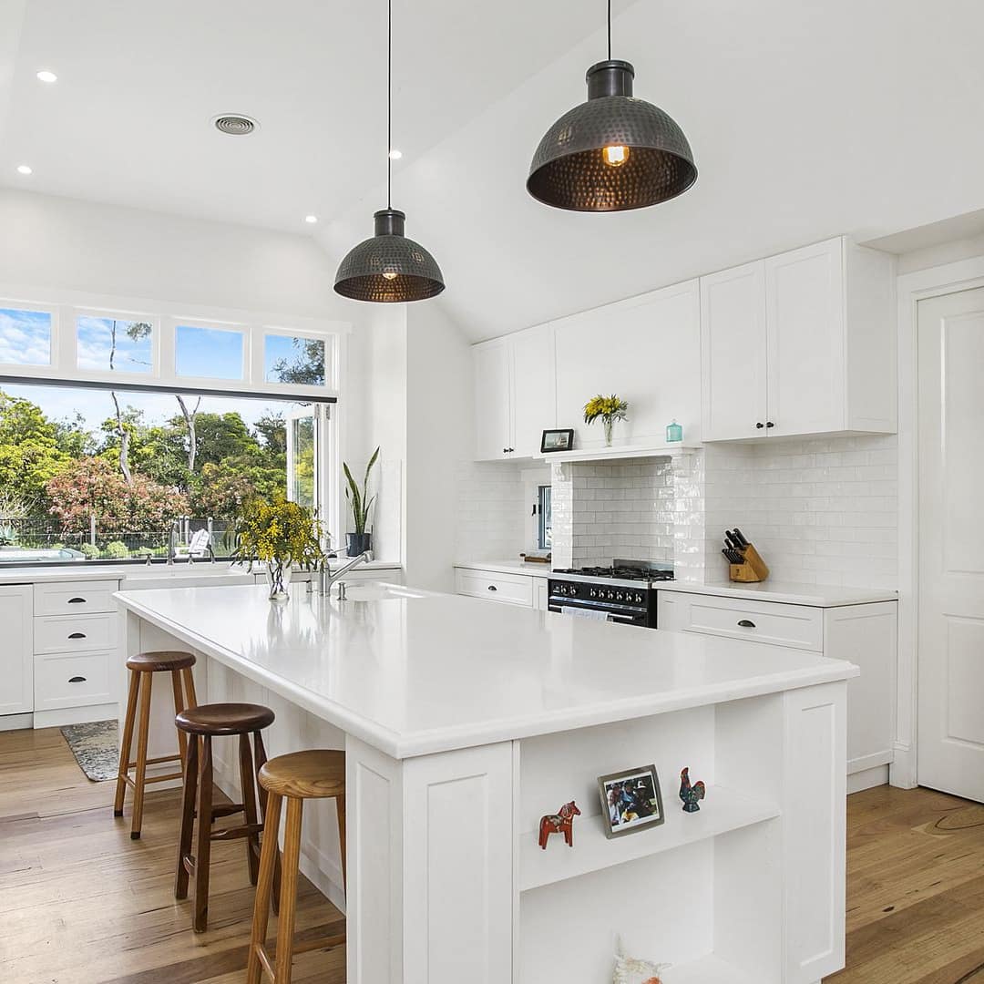 Farmhouse-style kitchen with white cabinets and countertops, island with wood stool, wide window with nature views.