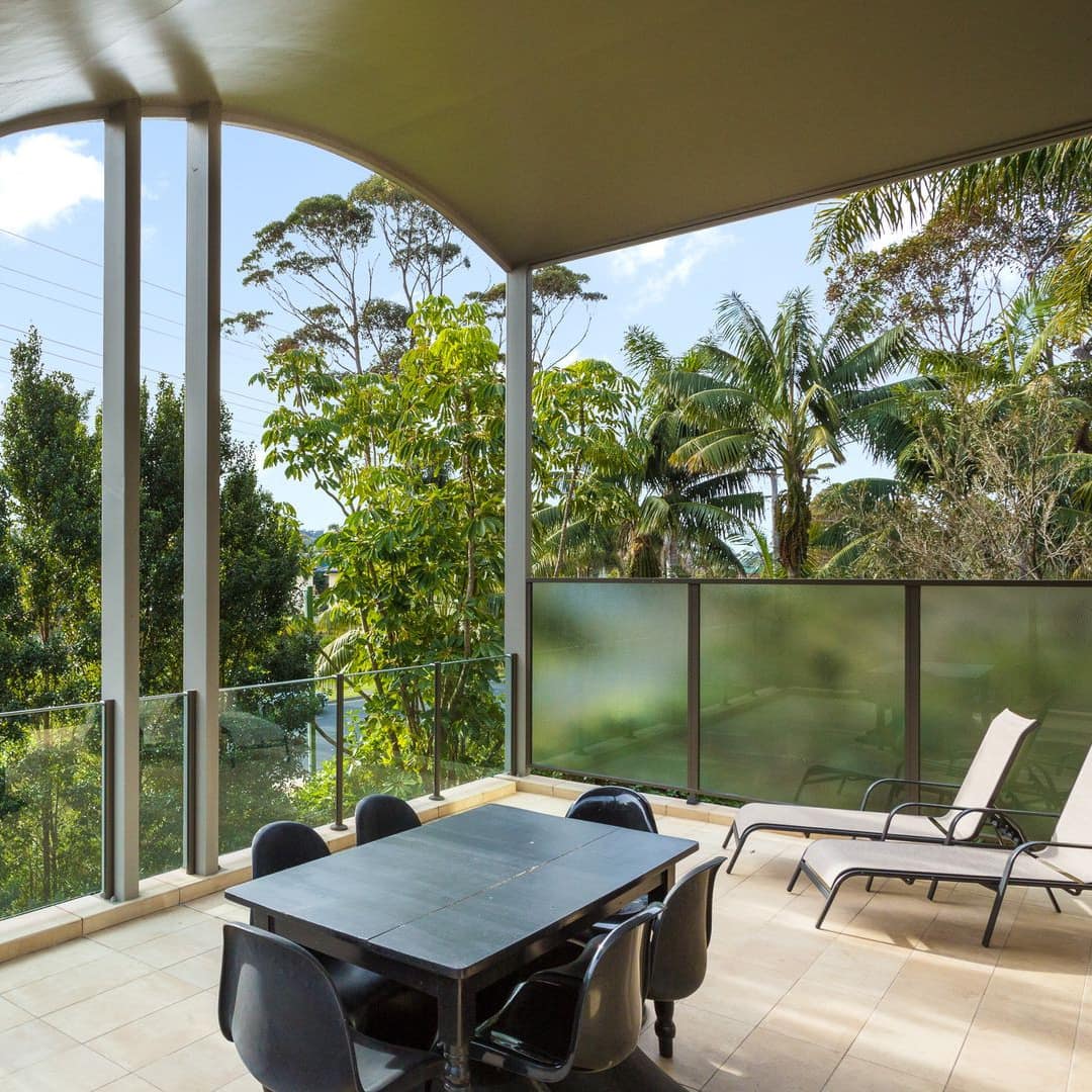 A large covered deck surrounded by trees, with glass railings, tan tile floor, white lounge chairs, black table and chairs.